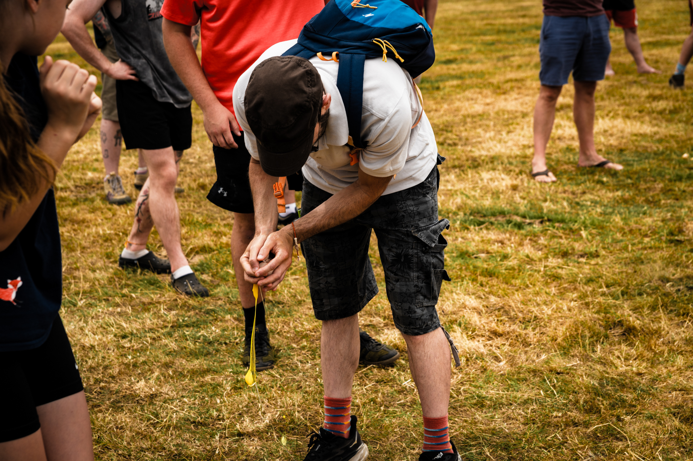 Boy in white t-shirt and dark shorts bending forward on grass field, adjusting something at his feet whilst other children stand nearby.