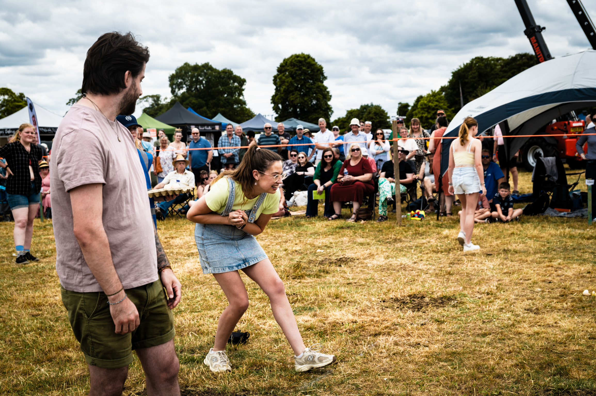 Woman in denim shorts pulls rope in tug-of-war game at outdoor festival, with man and crowd watching on grass field.