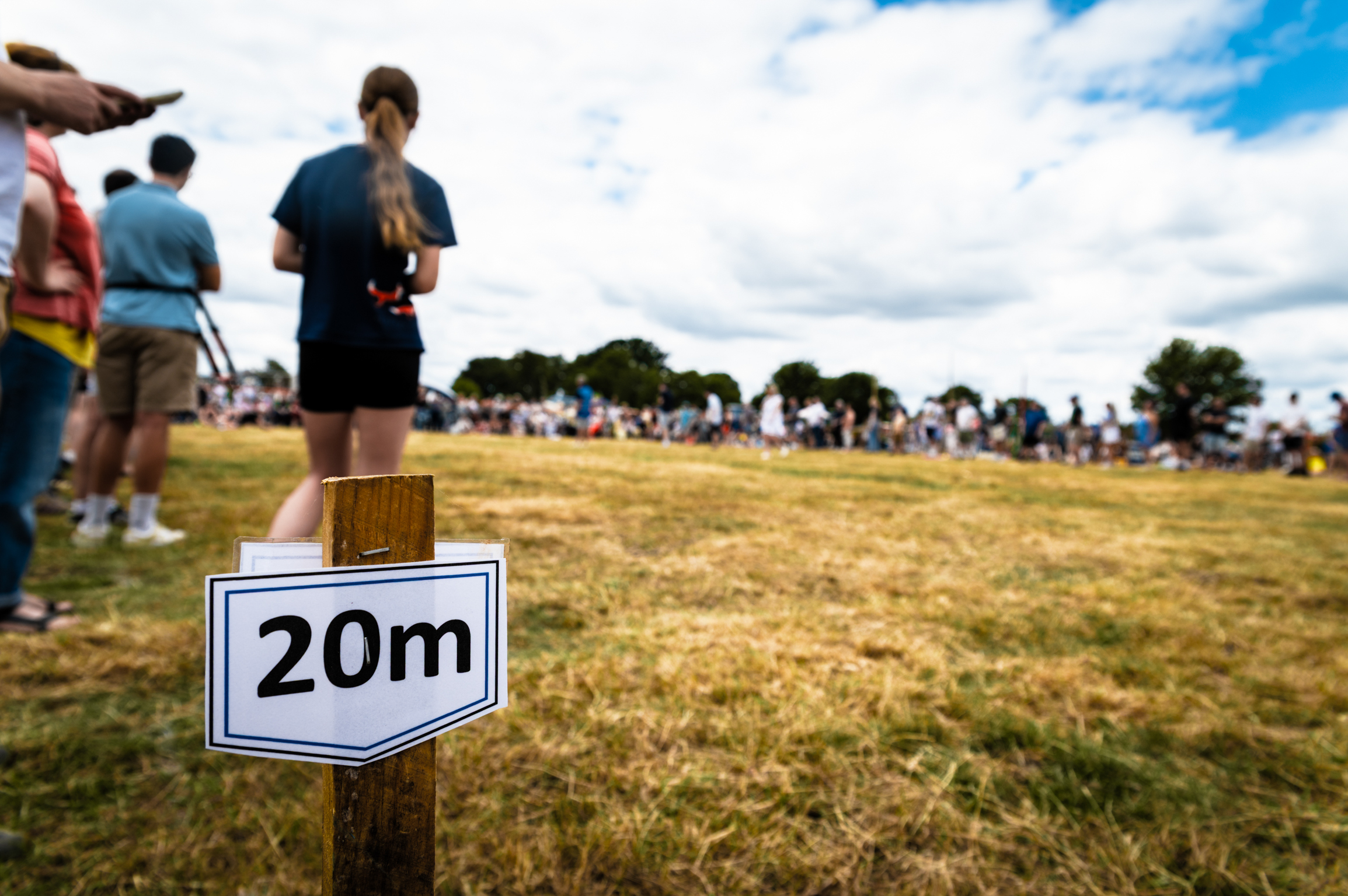 White distance marker sign reading "20m" on wooden post in foreground, people standing on grass field with trees and cloudy sky behind.