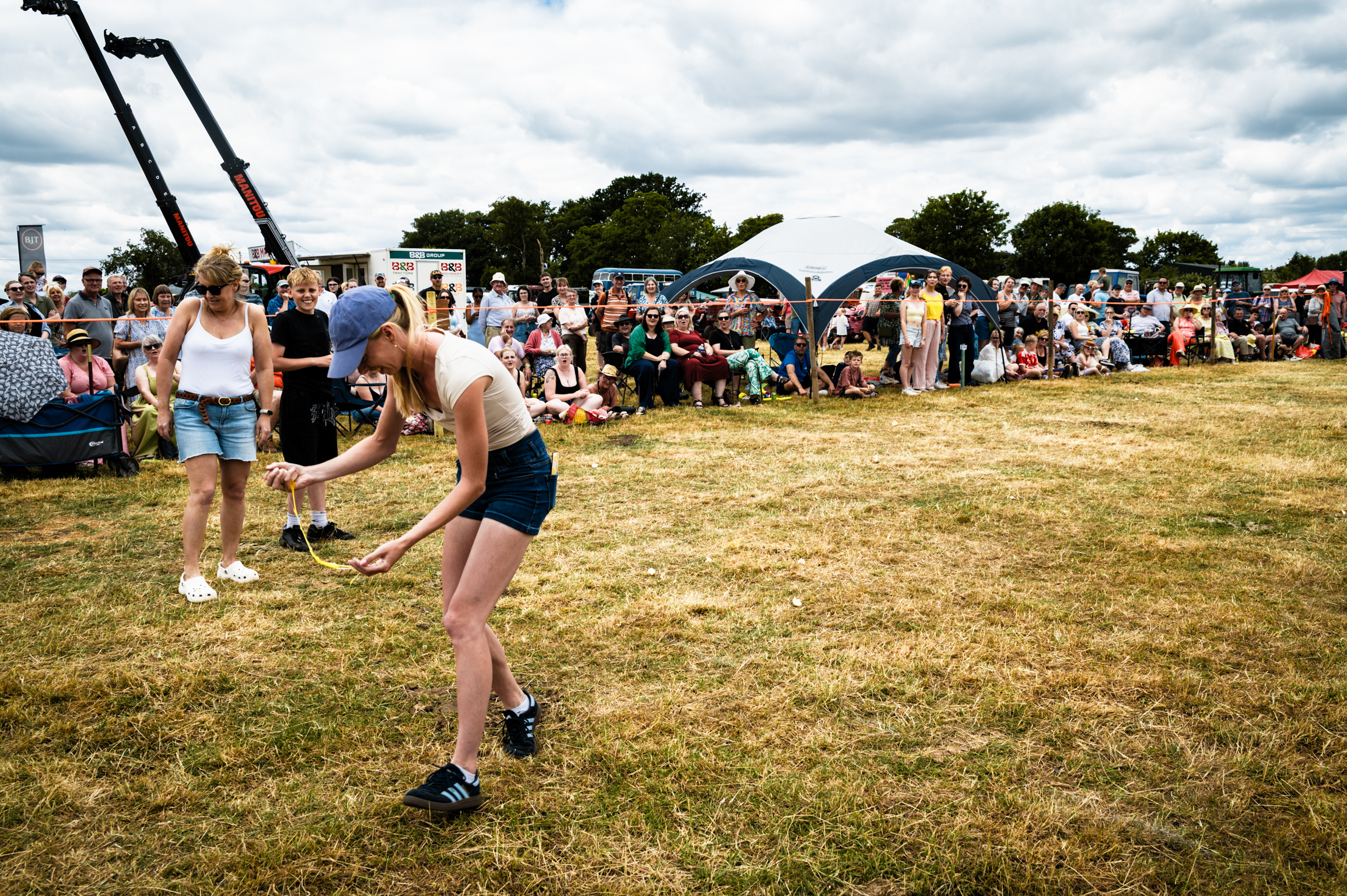 Woman in shorts and vest plays rounders on grass field at outdoor festival with tents and crowds in background under cloudy sky.