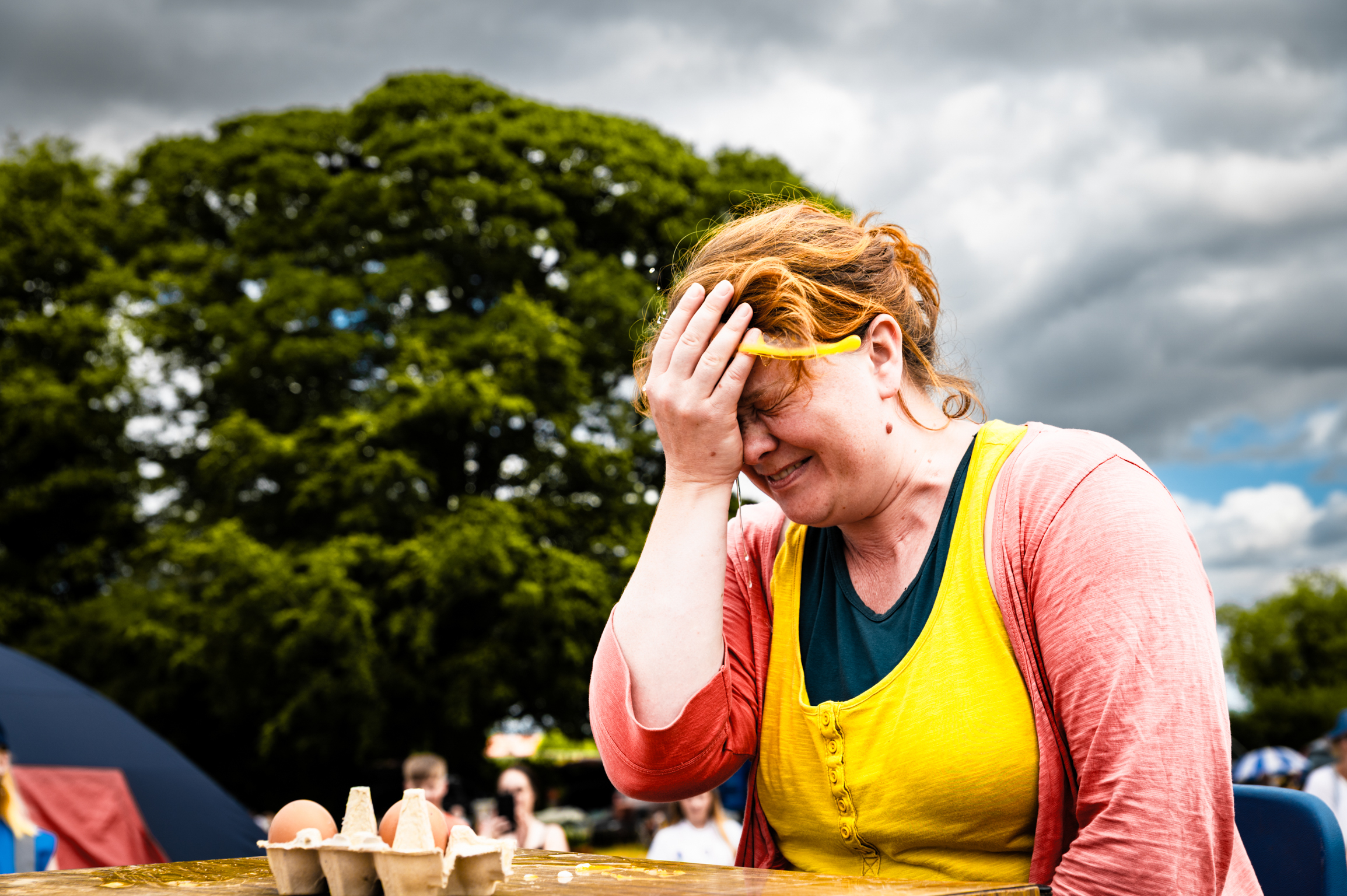 Woman in yellow top and pink cardigan with hand on forehead, seated outdoors with green trees and cloudy sky behind.