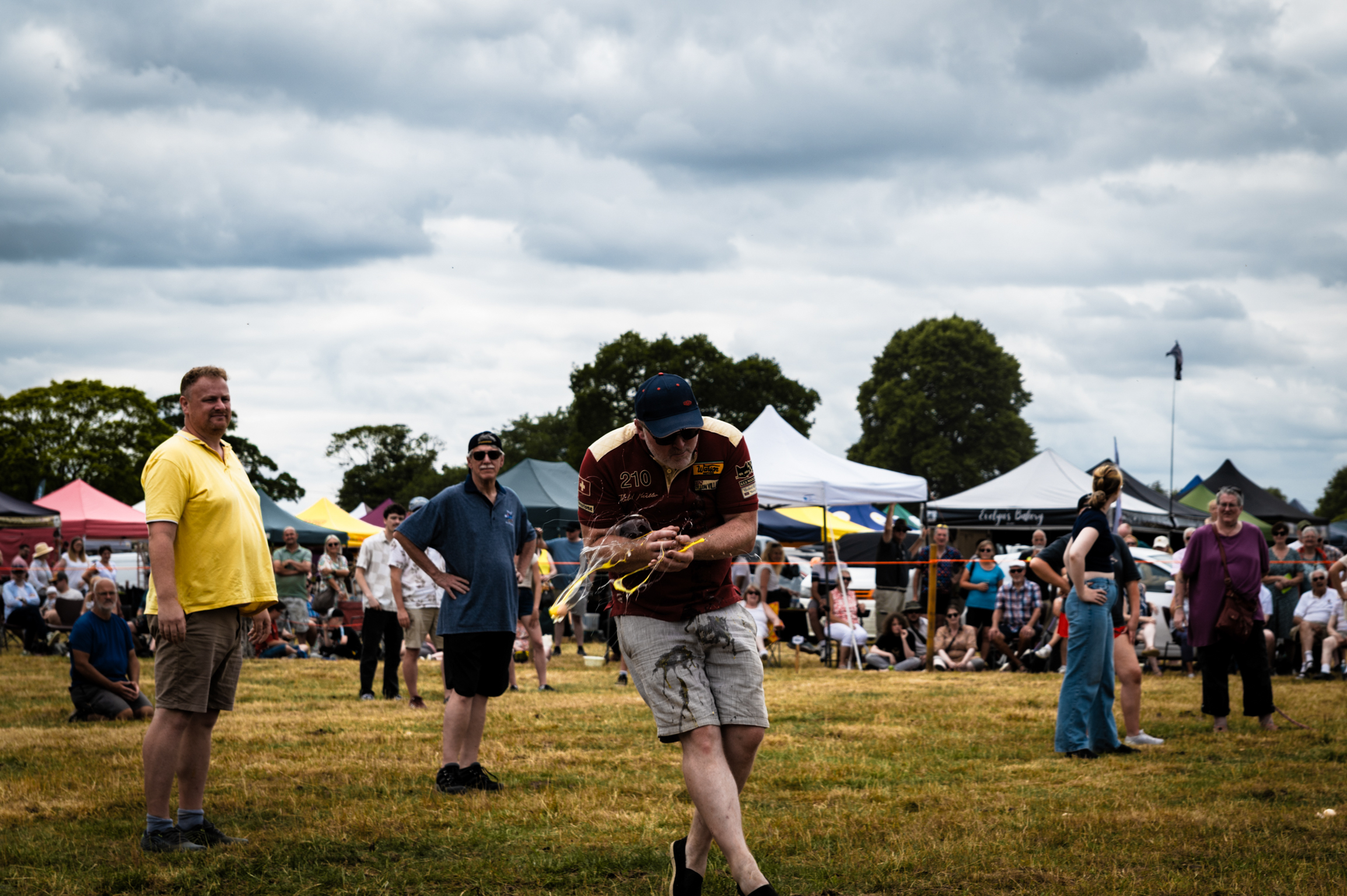 Tug of war competition at outdoor event with participants pulling rope, spectators watching under colourful gazebos, cloudy sky.