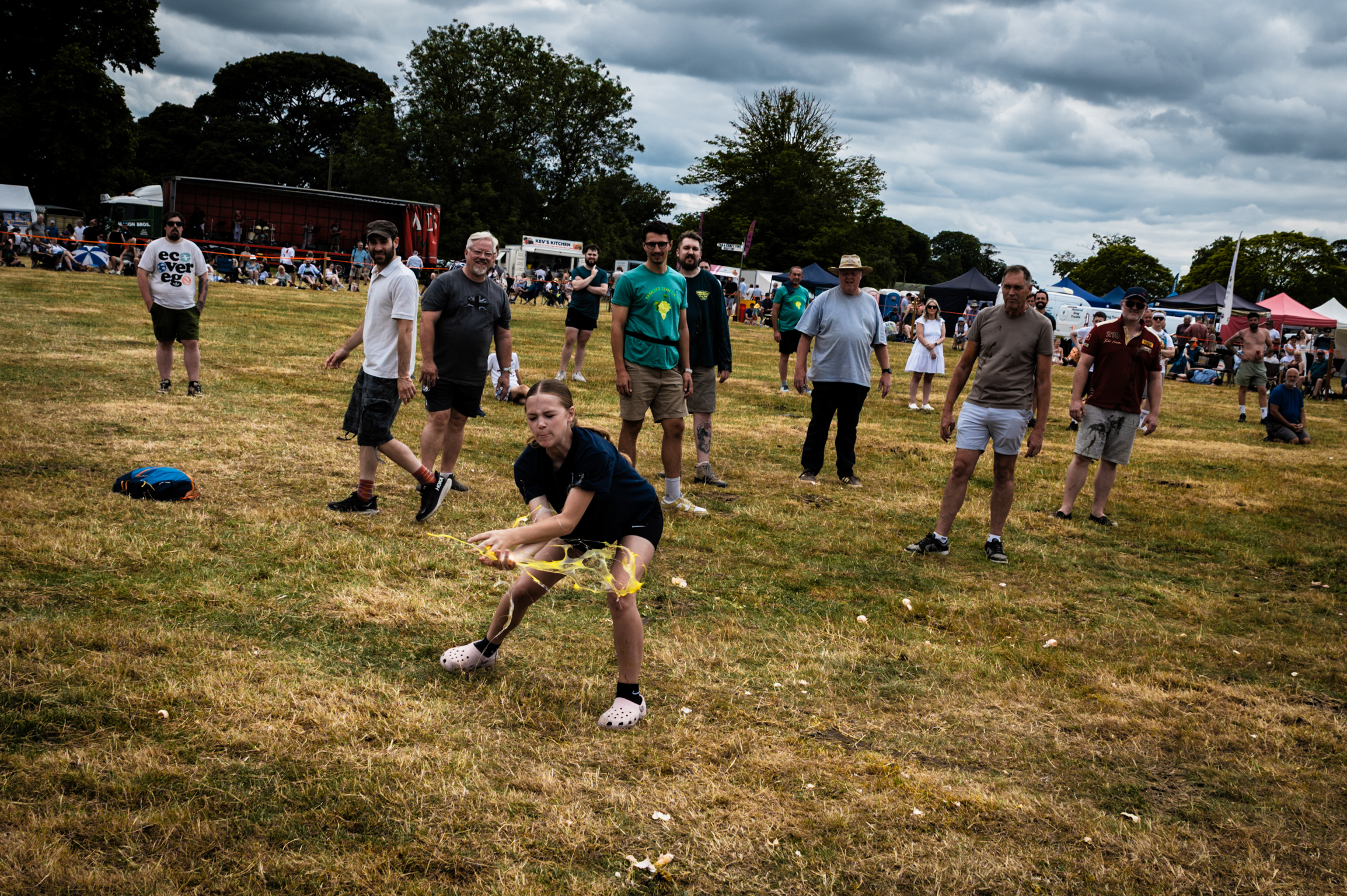 Man in black shirt pulls rope during tug-of-war on grass field, with spectators and trees in background under cloudy sky.