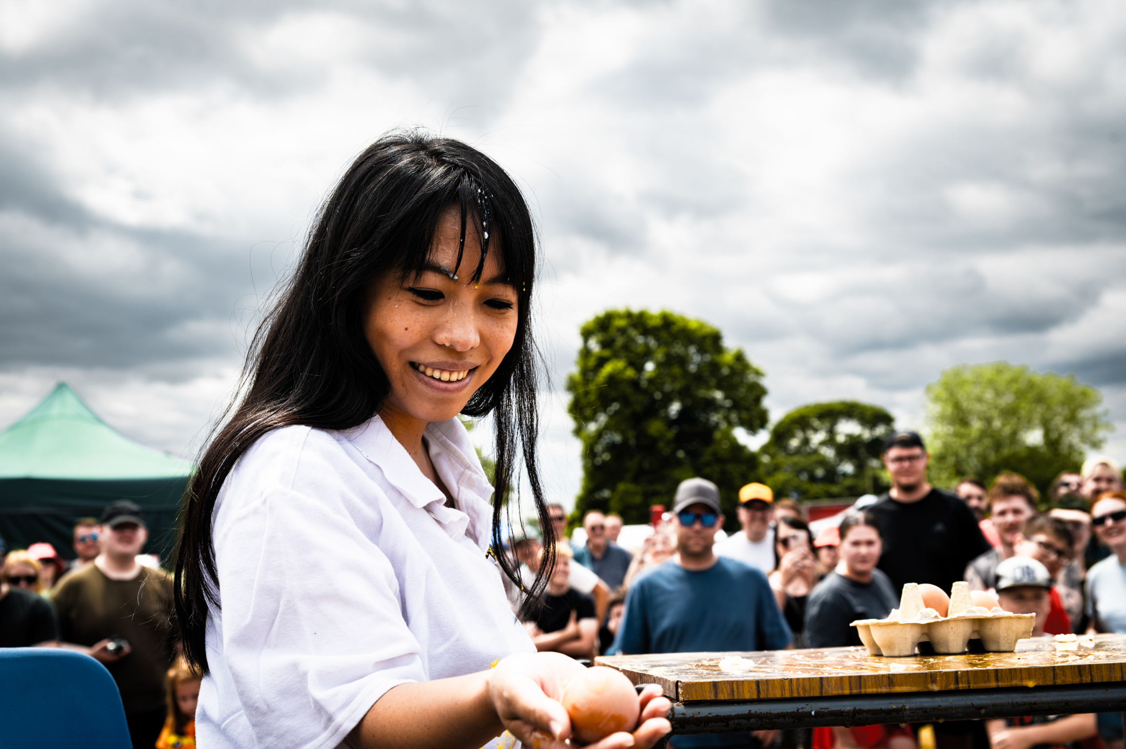 Woman with long dark hair in white shirt smiling whilst cooking at outdoor event with crowd and green trees in background.