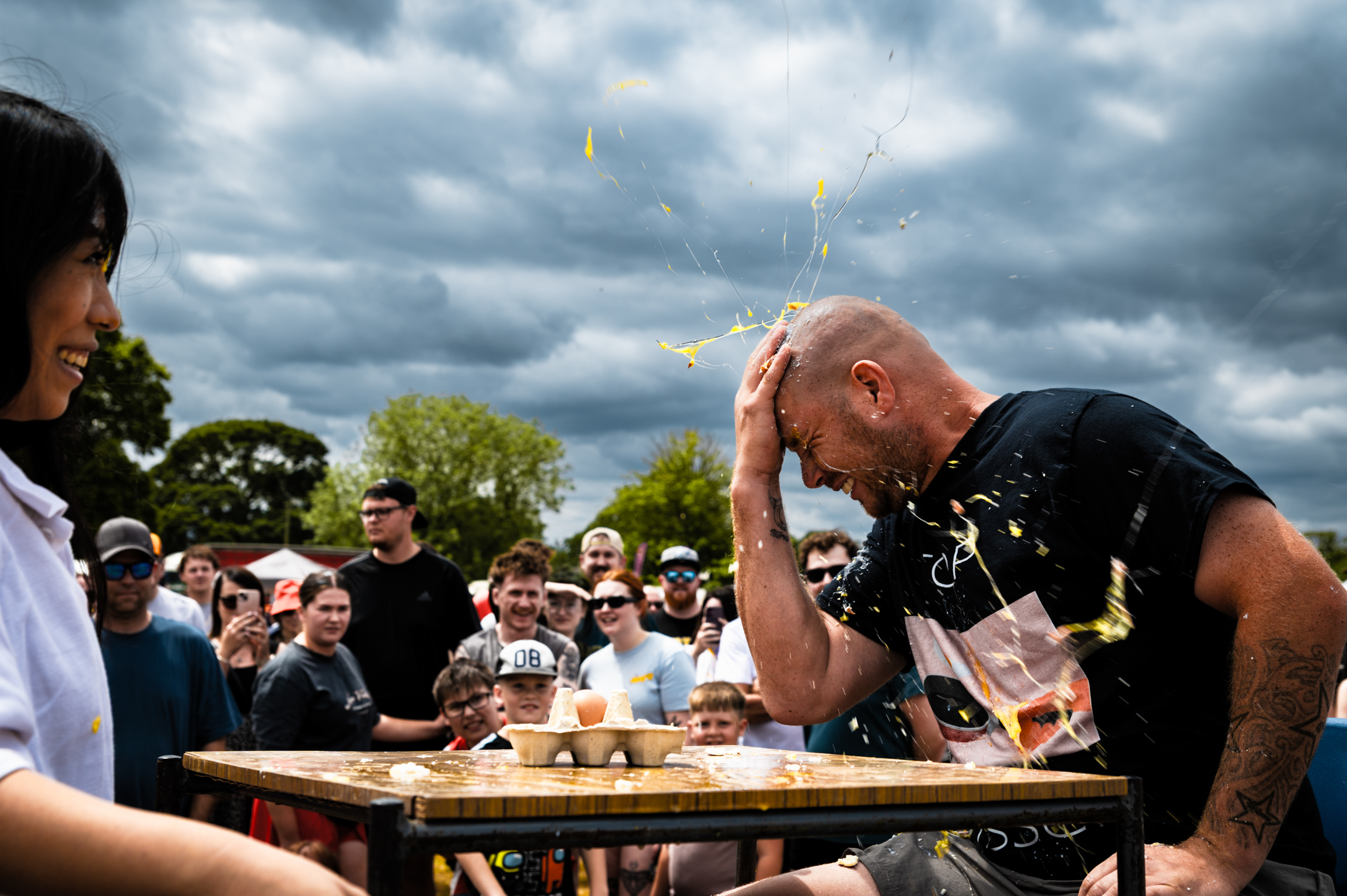 Man in black shirt at wooden table covered in yellow liquid, crowd watching, cloudy sky background.