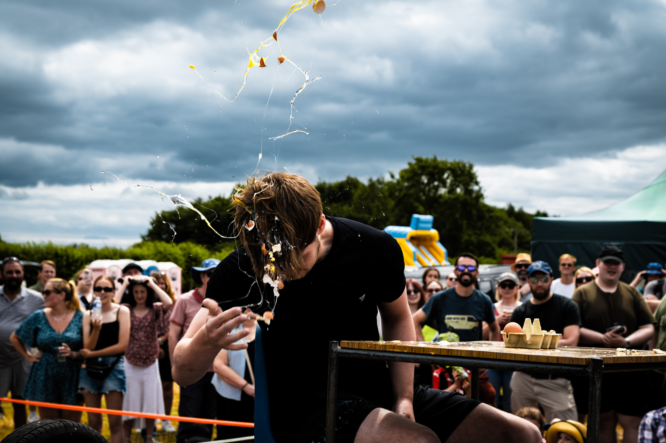 Man in black shirt eating at outdoor event with crowd watching under cloudy sky, yellow kite flying above.