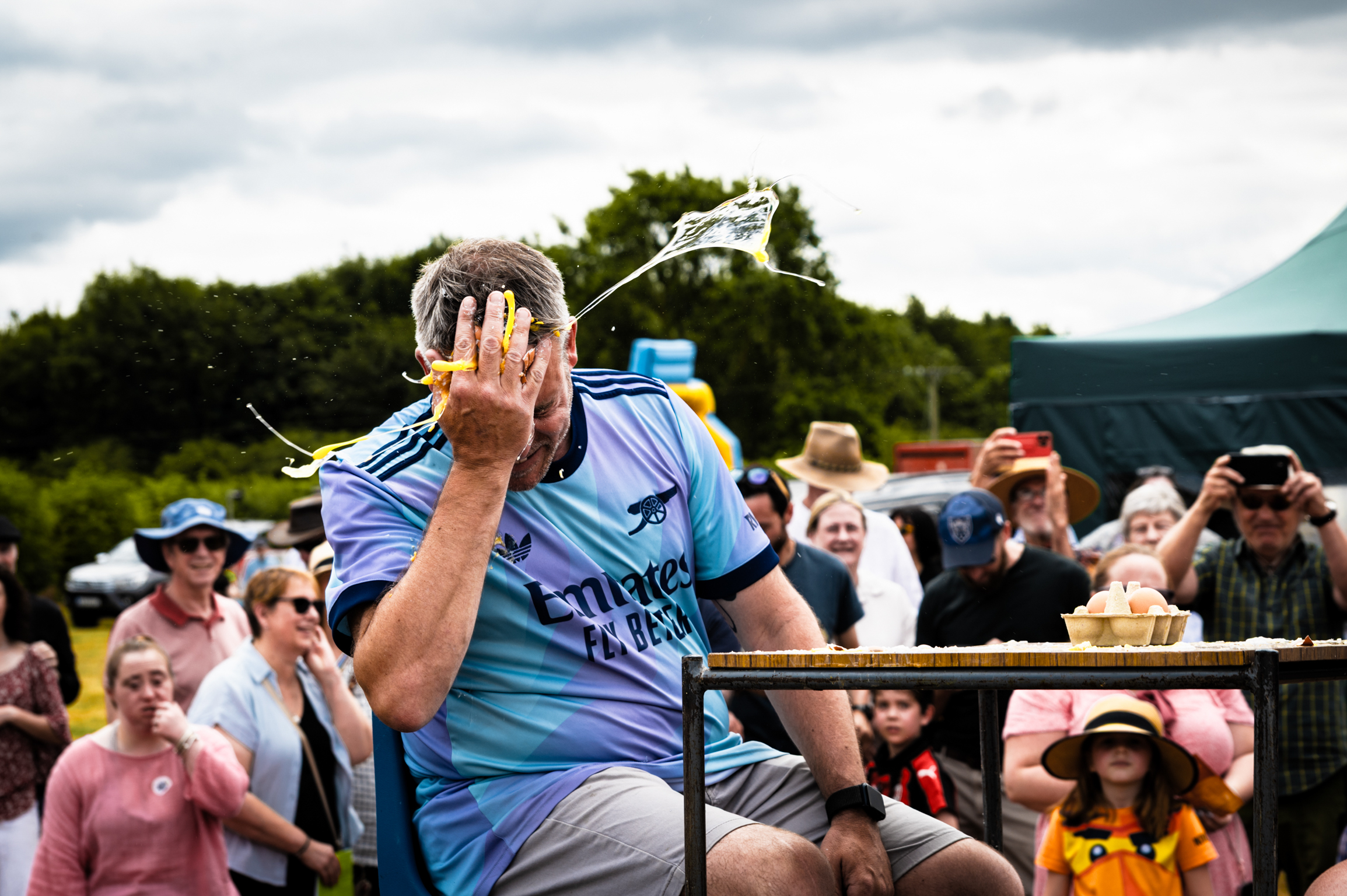 Man in blue sports shirt covers face with hand whilst sitting on stage, crowd of spectators watching in background under cloudy sky.
