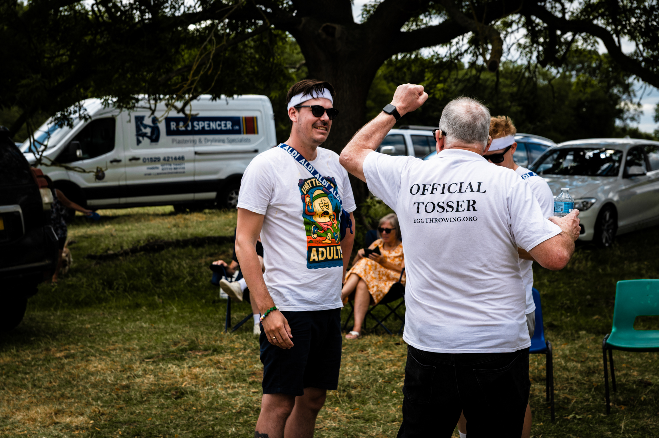 Two men outdoors, one wearing sunglasses and graphic t-shirt, other in "Official Tosser" shirt. Caravans and cars in background under trees.