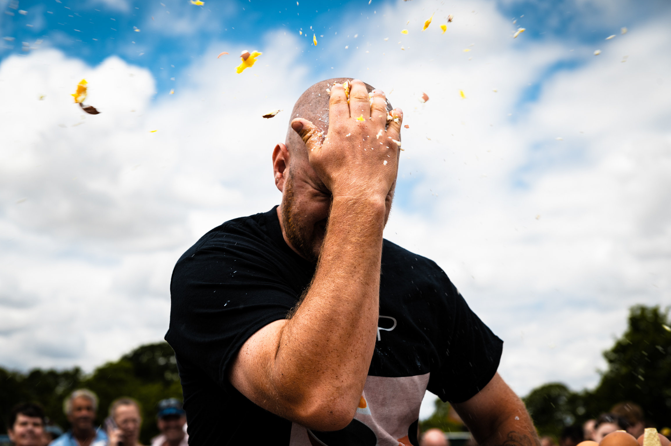 Bald man in black shirt covering face with hand, confetti falling from blue sky with white clouds, crowd in background.