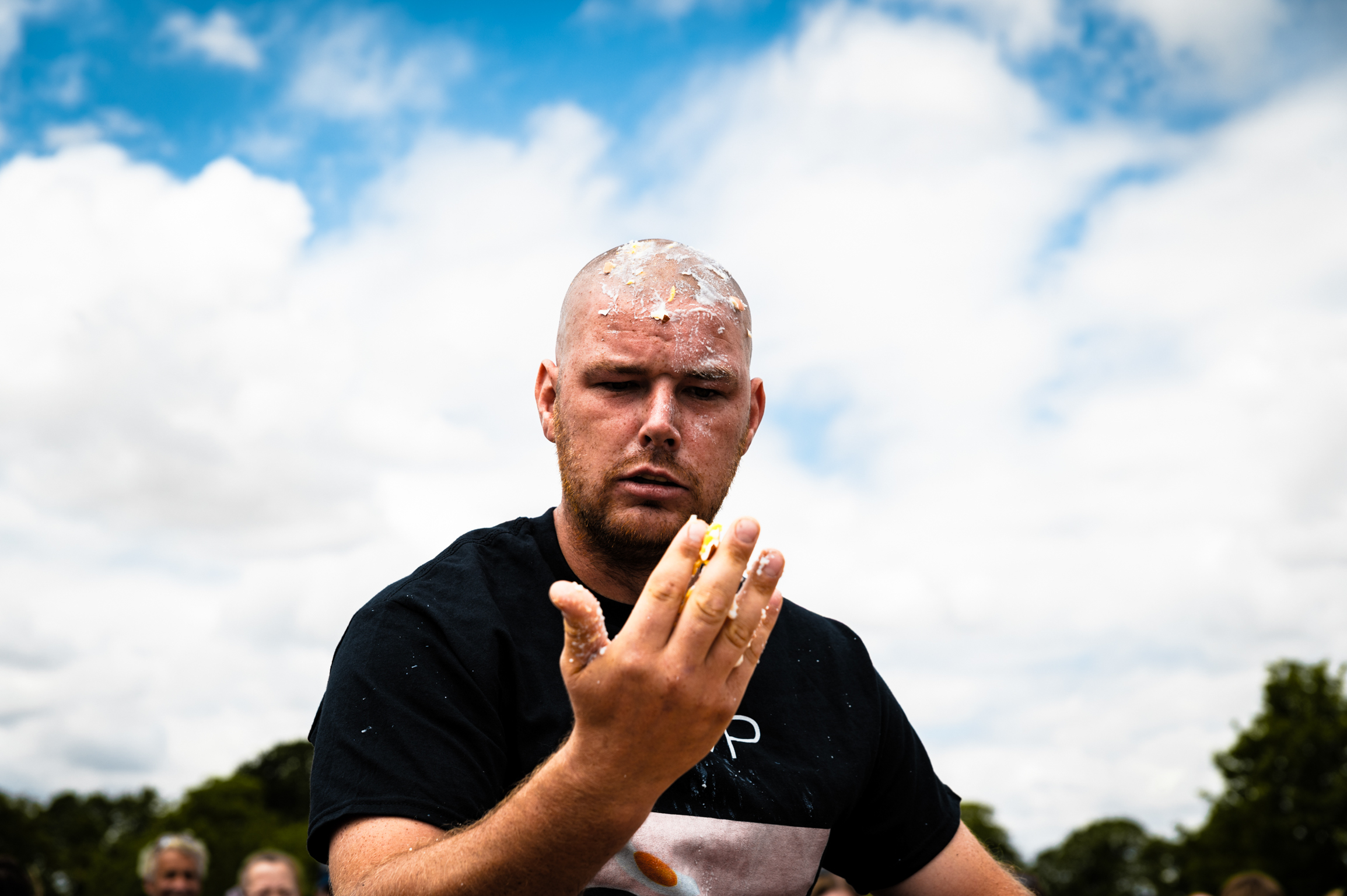 Bald man in black shirt holds up hand towards camera against blue sky with white clouds and green trees in background.