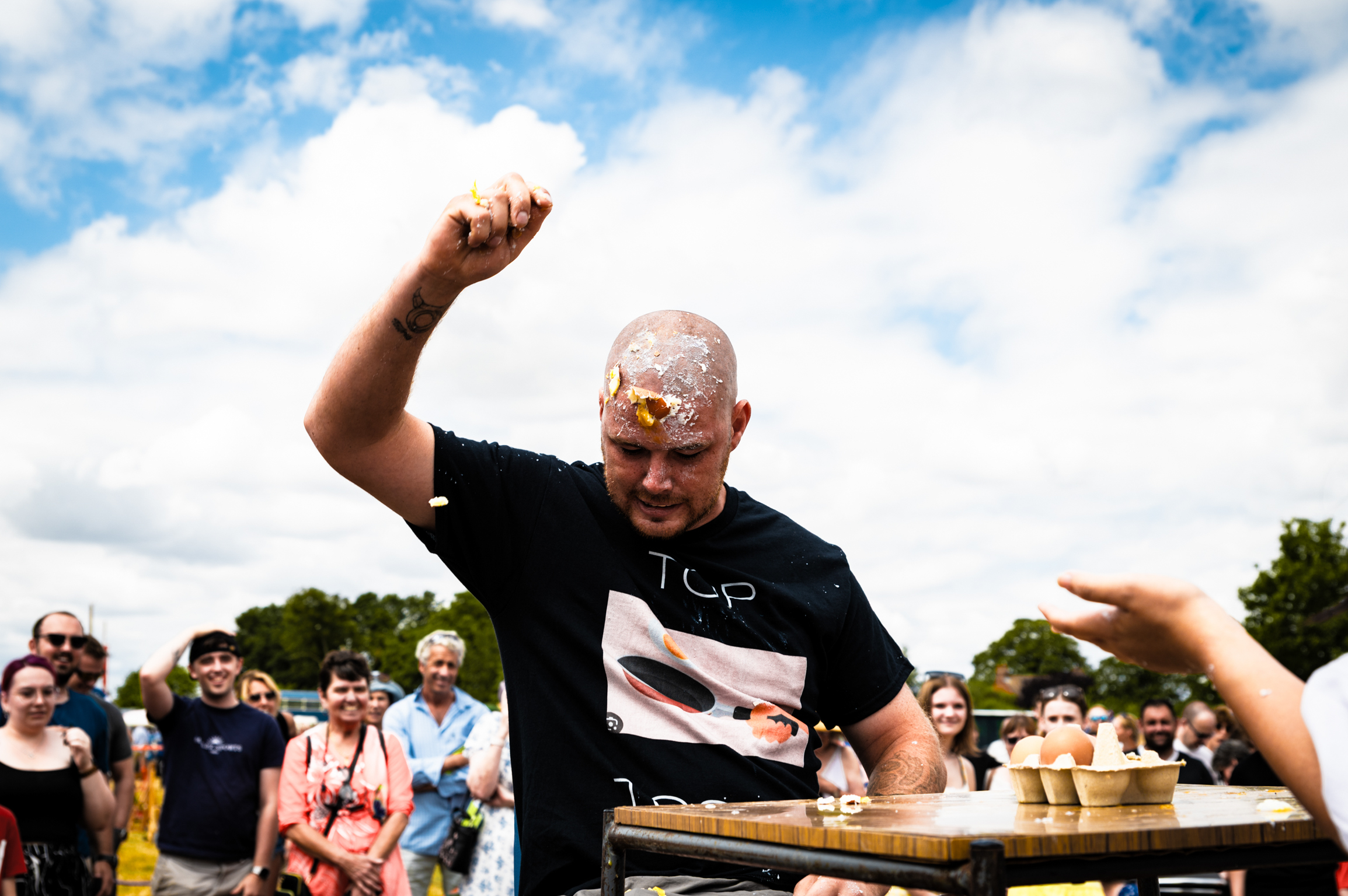 Bald man in black t-shirt raises fist in celebration at outdoor event with crowd and blue sky with white clouds behind him.