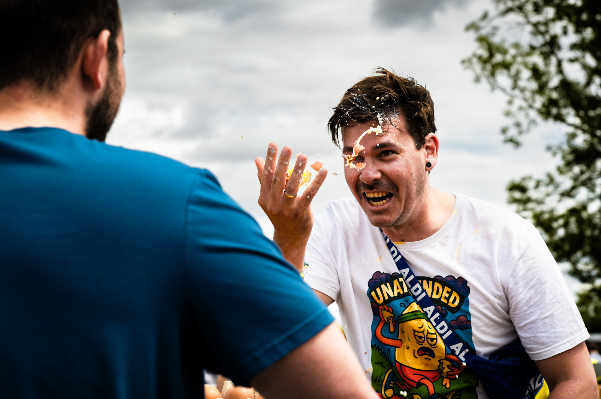 Man in white graphic t-shirt giving high-five to person in blue shirt outdoors, trees and cloudy sky in background.