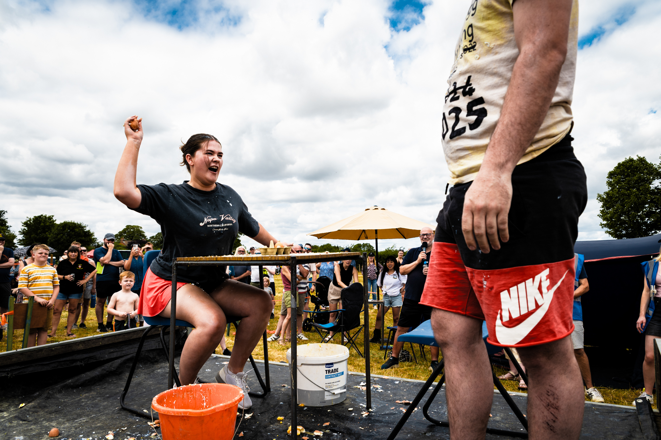 Two people playing table tennis outdoors at community event, one celebrating with raised fist, crowd watching in background.