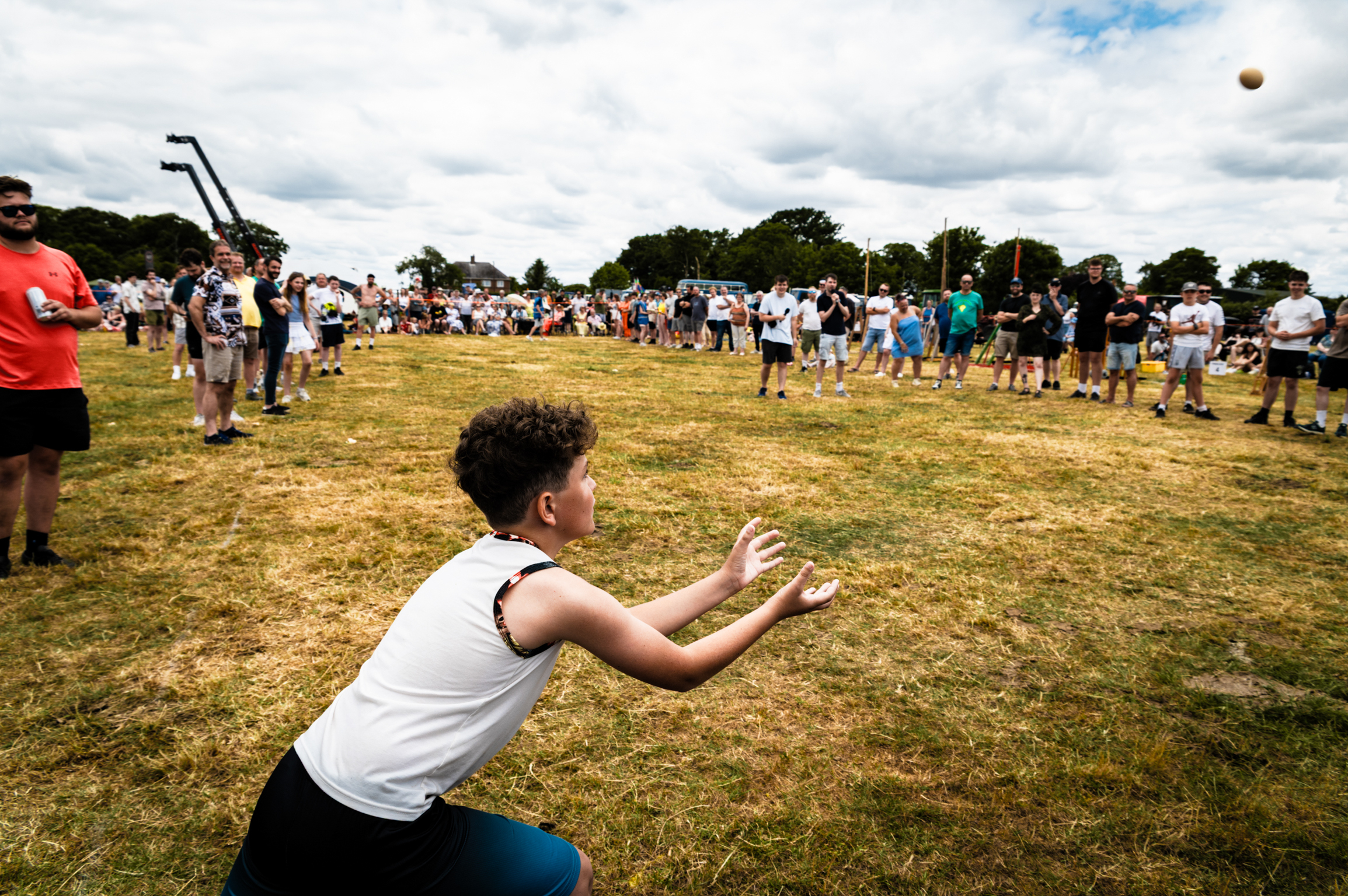 Woman in white vest catching ball on grassy field with crowd watching. Cloudy sky with trees in background.