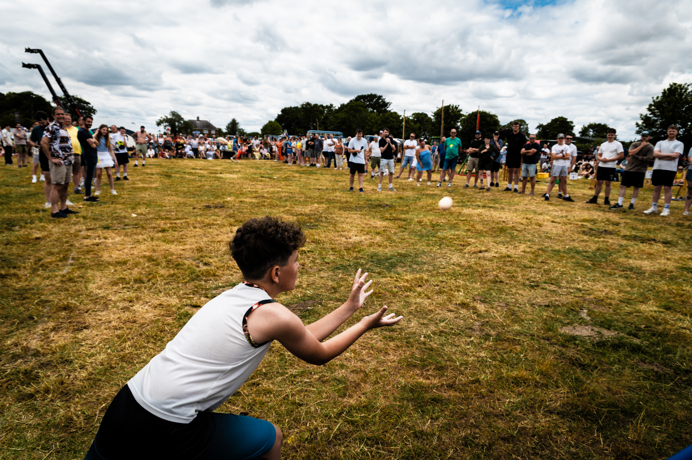 Person in white vest crouching on grass field with crowd watching under cloudy sky at outdoor sports event.