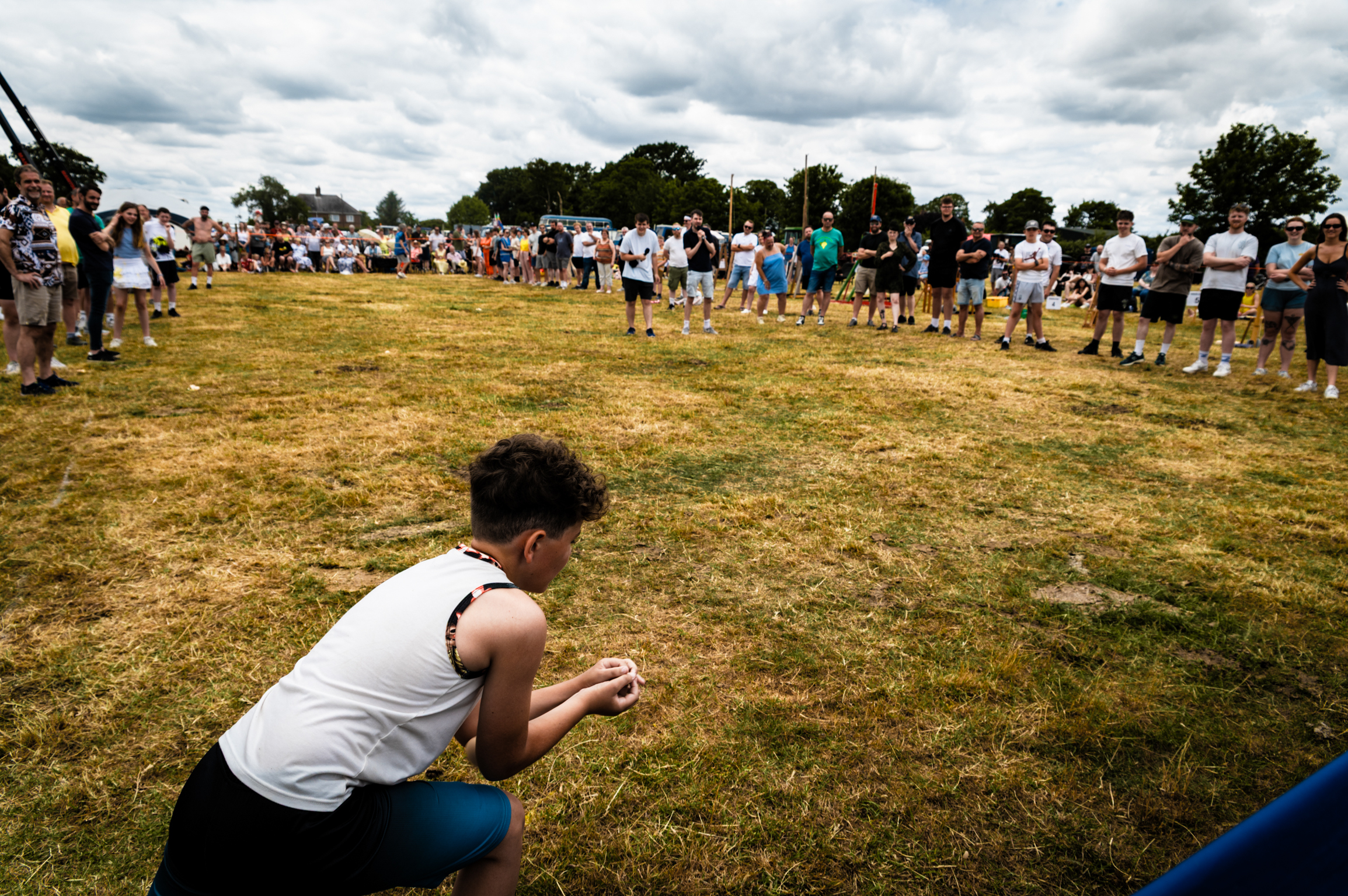 Person in white vest crouching on grass field with crowd forming circle around perimeter, trees and cloudy sky in background.