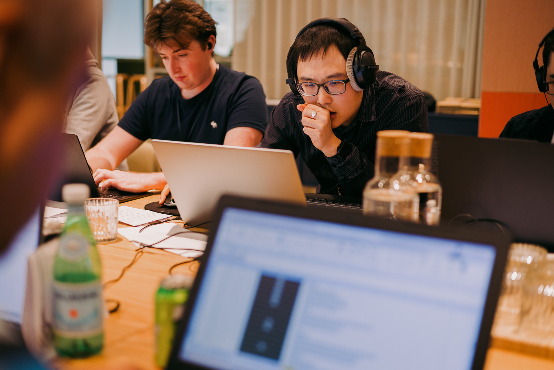 Two men working at laptops in office setting, one wearing headphones and glasses, wooden desk with bottles and papers visible.