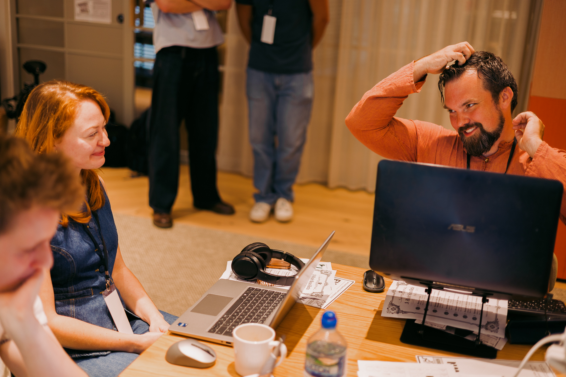 Man in orange jumper with headphones at desk with laptop, woman in dark top seated nearby, people standing in background.