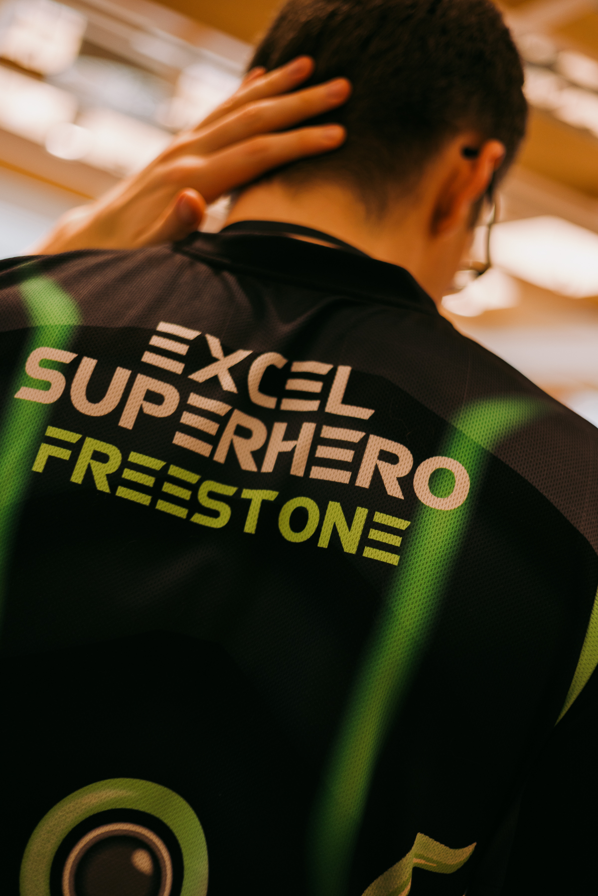Person wearing black jacket with "EXCEL SUPERHERO FREESTONE" text in green and yellow letters, hand on head, wooden floor background.