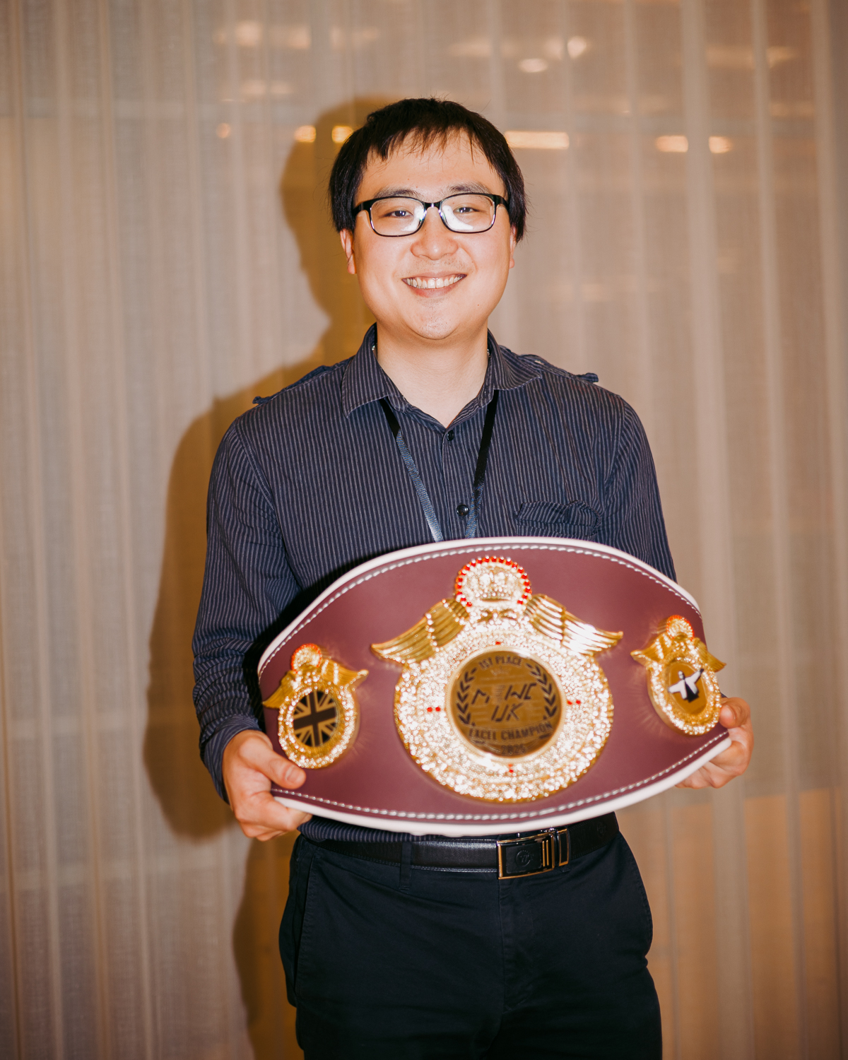 Person wearing glasses and dark striped shirt holding large maroon and gold championship belt with ornate decorations.