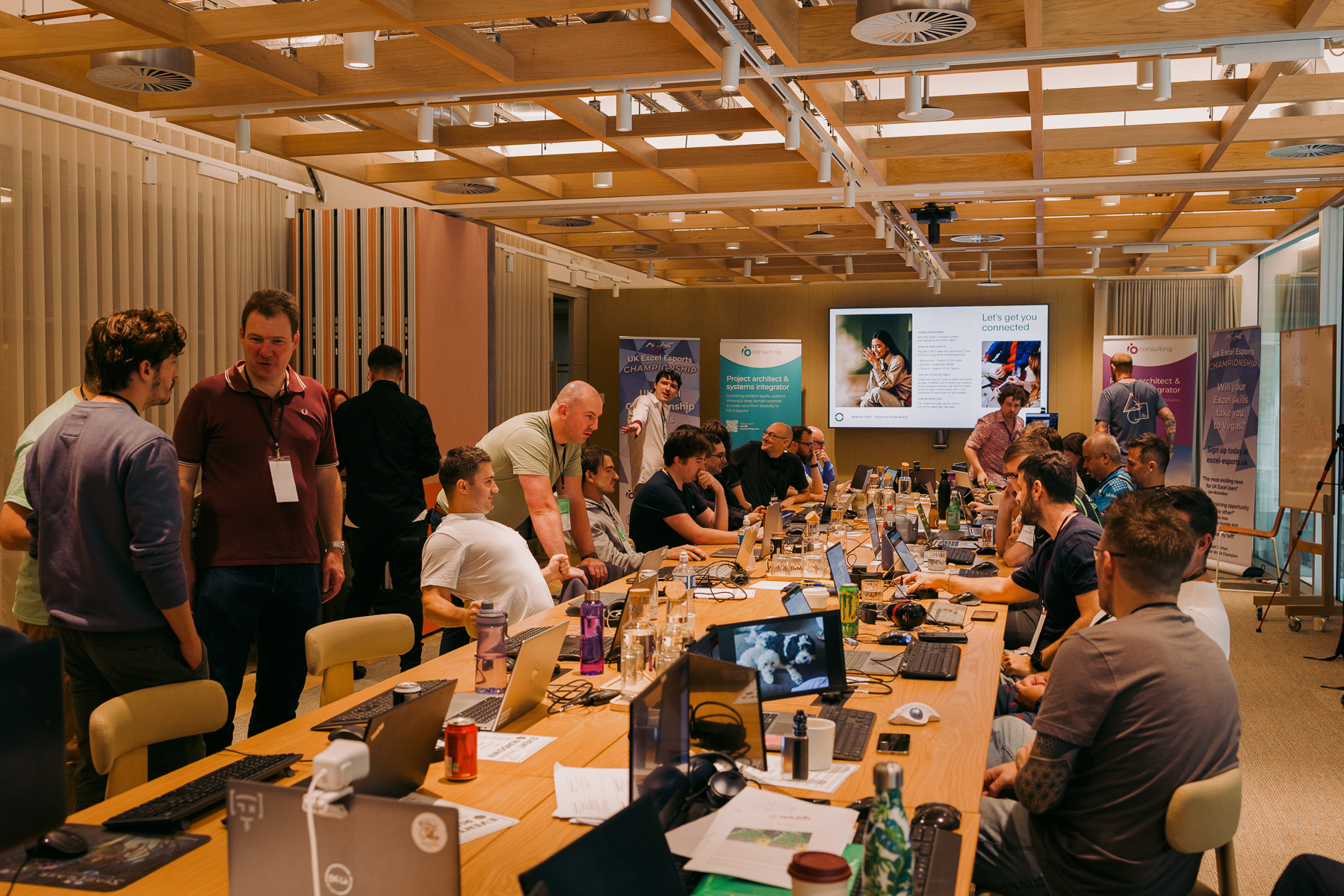 Conference room with people seated around long tables, laptops and bottles visible, wood-panelled ceiling, presentation screens on wall.
