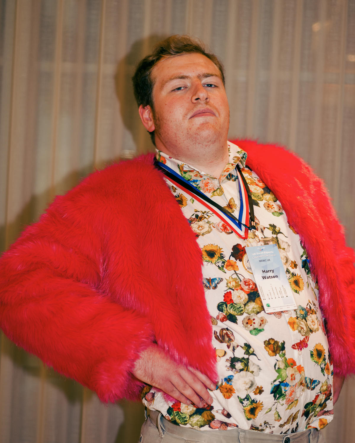 Man wearing bright red feathered cape over floral patterned shirt with medal and name badge, posed with hand on hip.