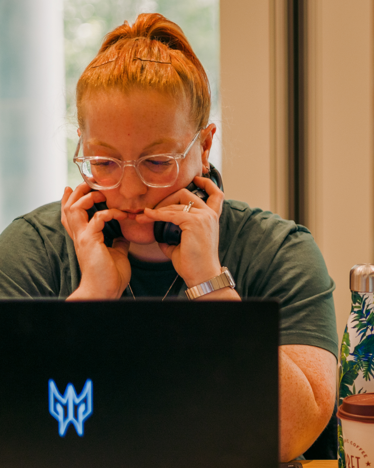 Person with ginger hair in bun, wearing glasses and olive green shirt, hands covering face whilst sitting at laptop with blue logo.