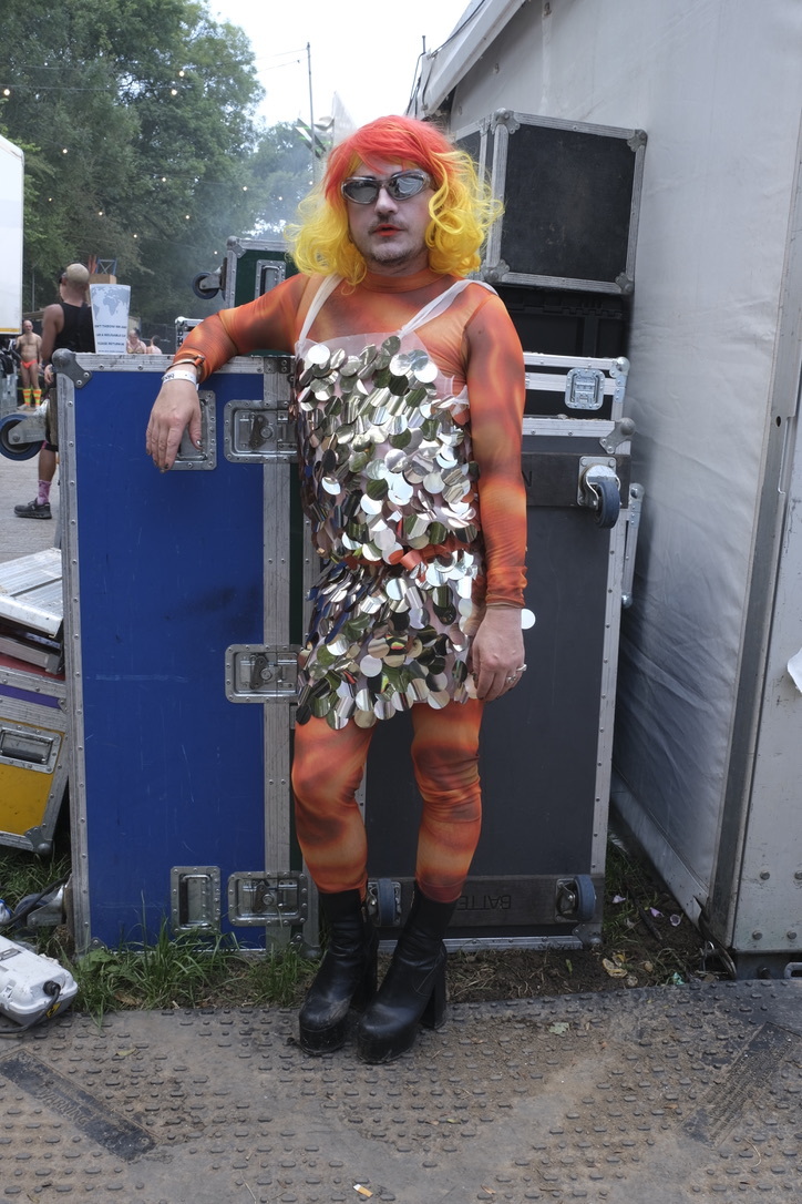 Person in drag with bright orange and yellow wig, sequined silver dress, orange tights, standing by blue equipment case and white trailer.