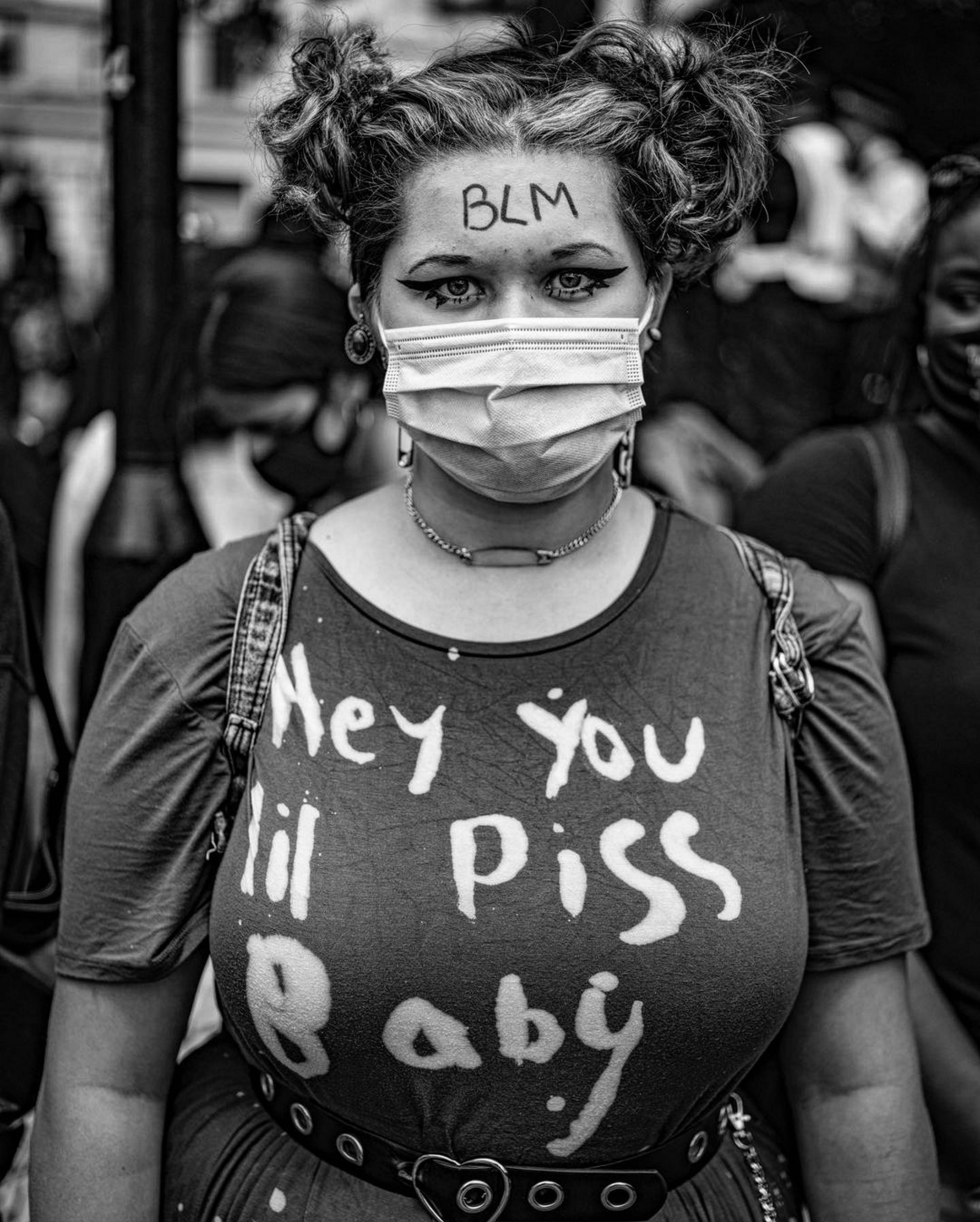 A woman wearing a protective face mask with the text "BLM" written on it, and a t-shirt with the text "Hey you lil piss baby" printed on it.