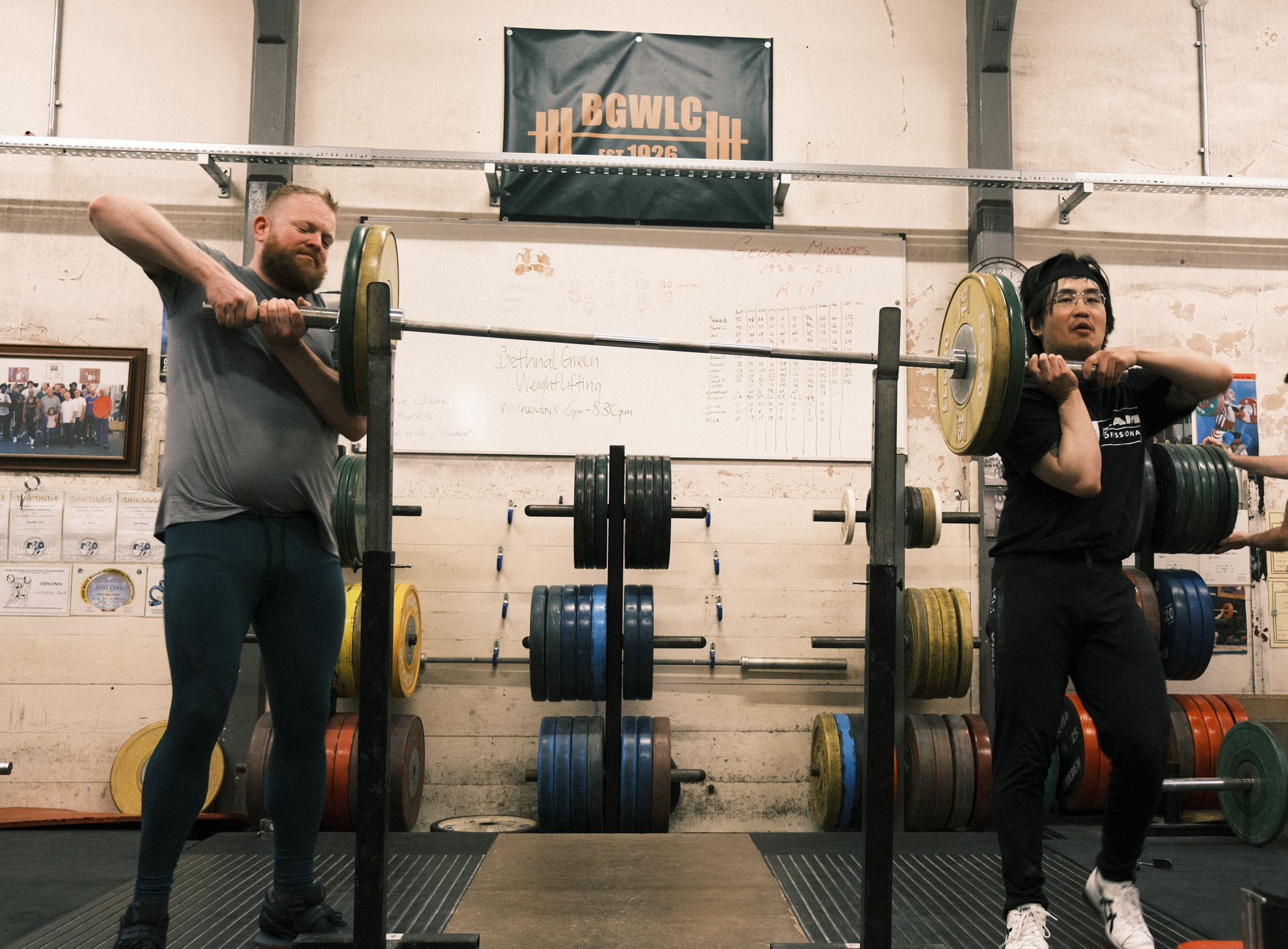 Two people weightlifting with barbells in gym, man on left stretching arm, woman on right holding barbell across shoulders.