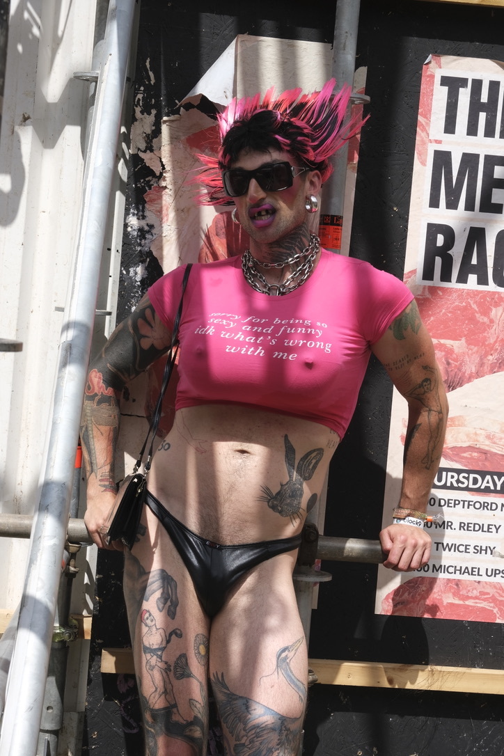 Person with bright pink hair wearing sunglasses, pink crop top, black underwear, and multiple tattoos standing outdoors near signage.