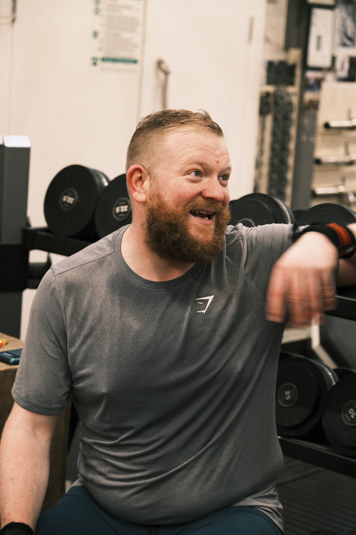 Bearded man in grey athletic shirt smiling whilst sitting on gym equipment, with dumbbells and exercise machines in background.