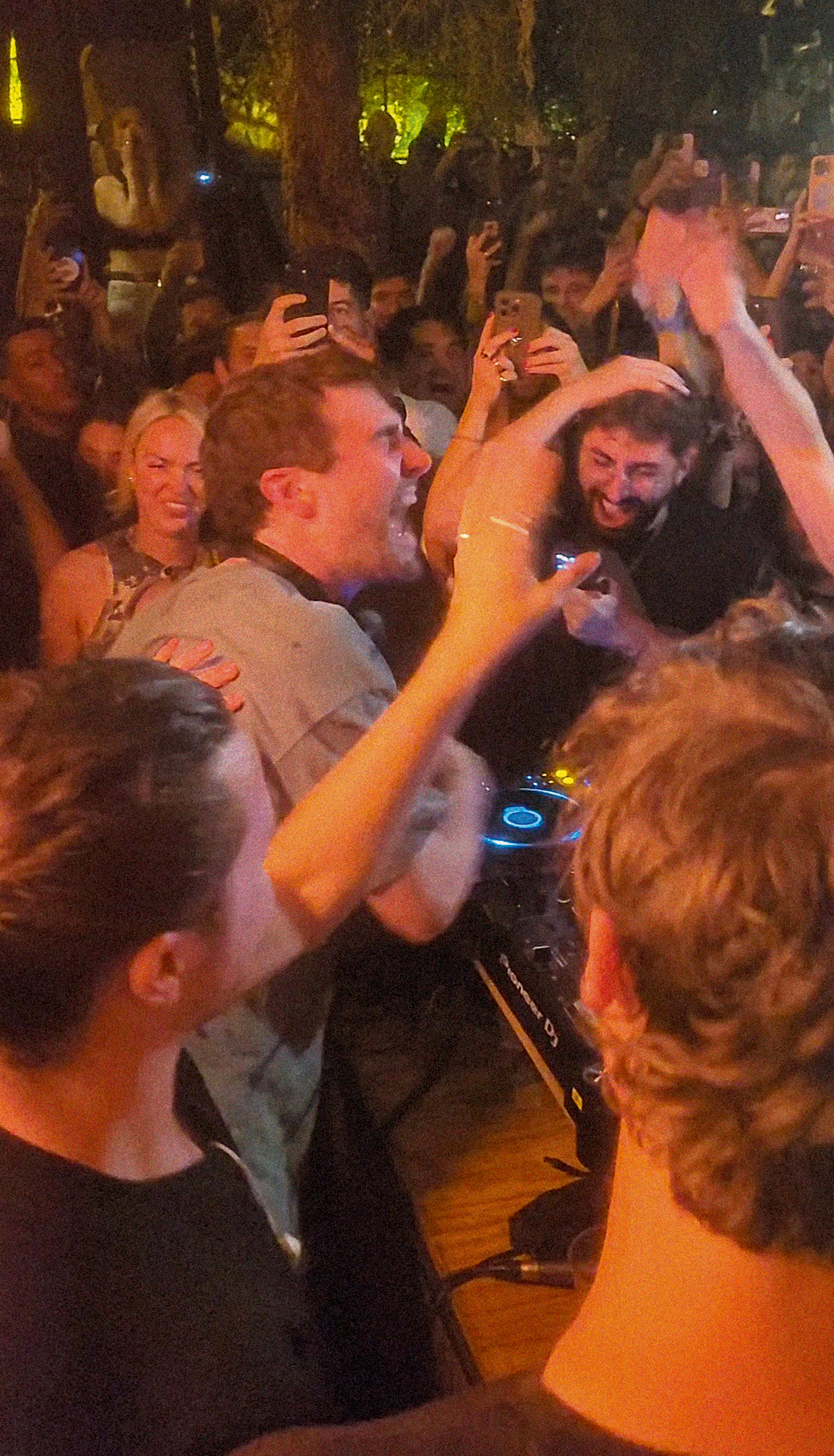 Crowded indoor venue with people raising mobile phones, warm orange lighting, green foliage visible above, faces illuminated by screens.