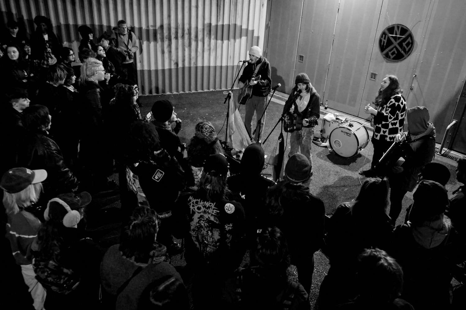 Black and white image of band performing on small stage with drum kit, guitars, surrounded by seated audience in intimate venue.