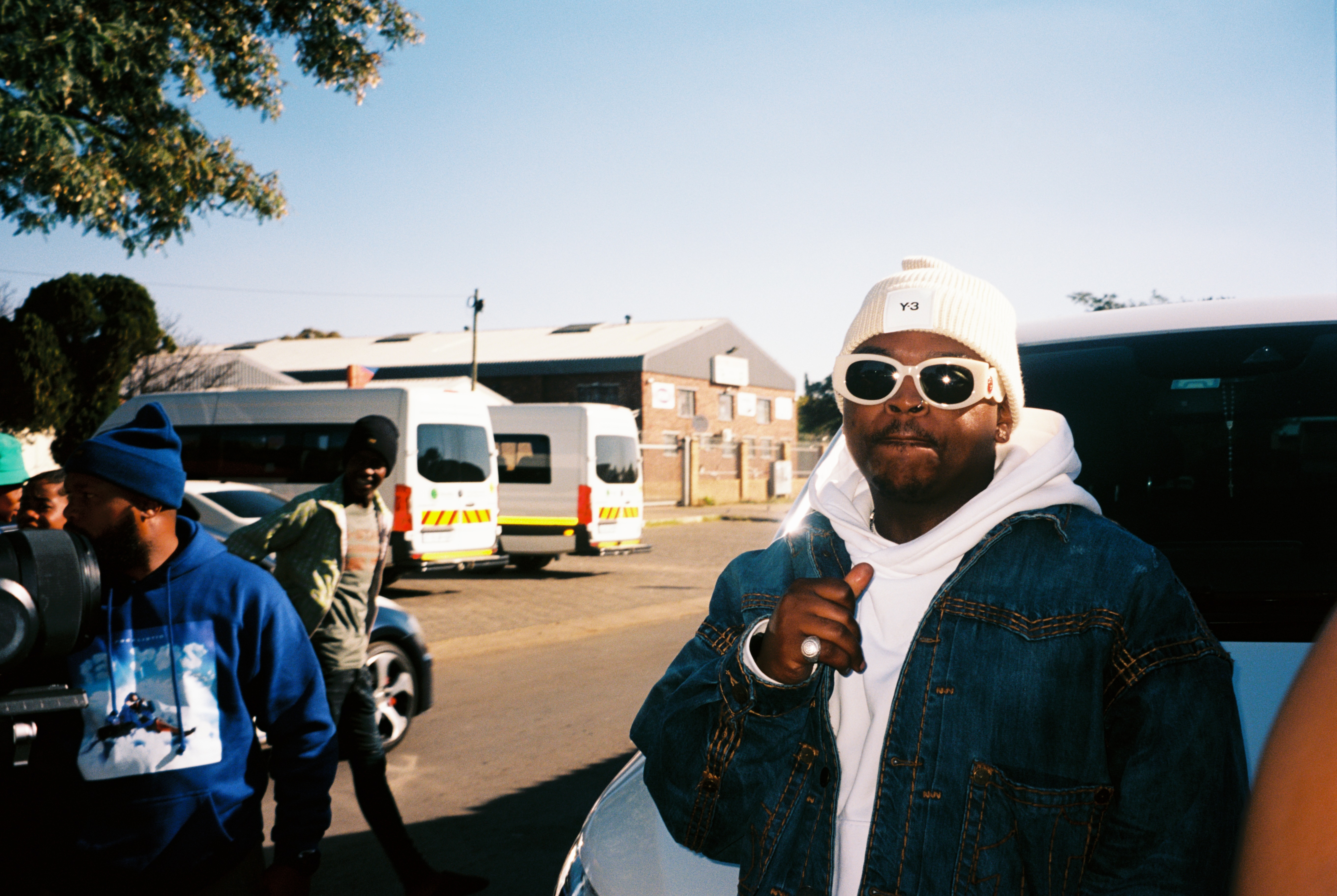 A bearded person in sunglasses and a hood stands in the foreground, with a crowd of people and vehicles in the background.