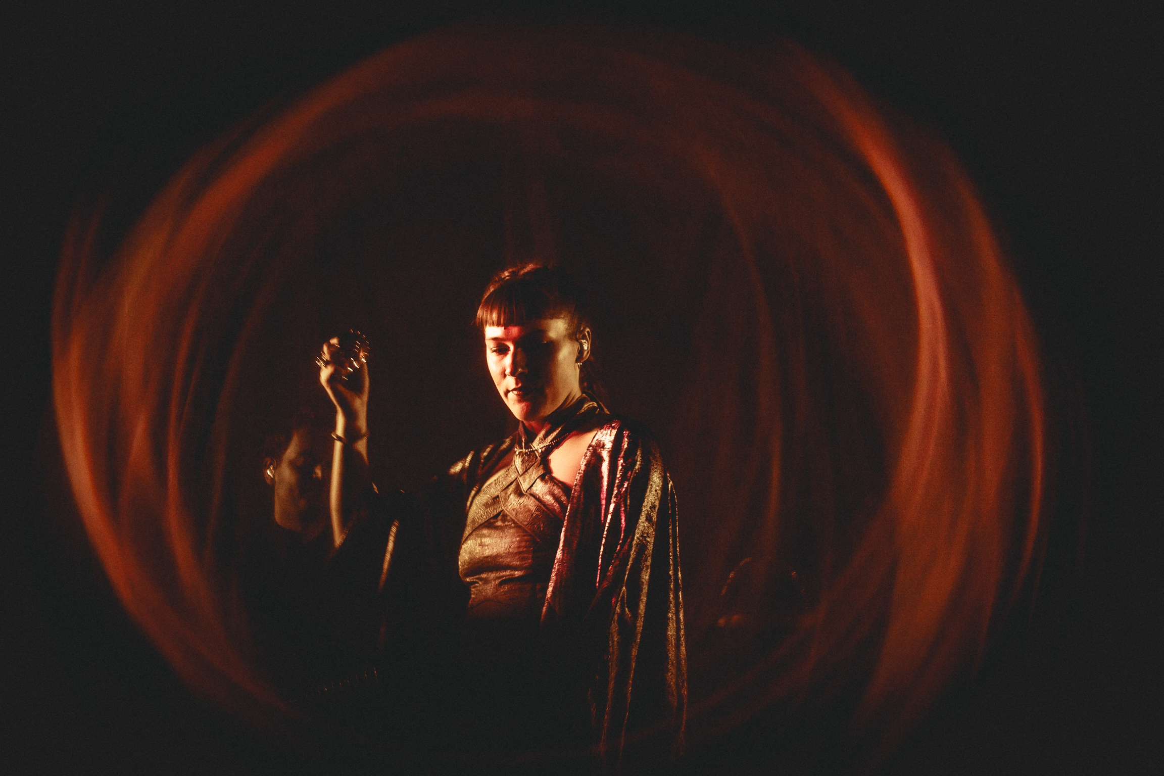 Man in dark clothing stands centre frame, illuminated by warm orange circular light patterns against black background.