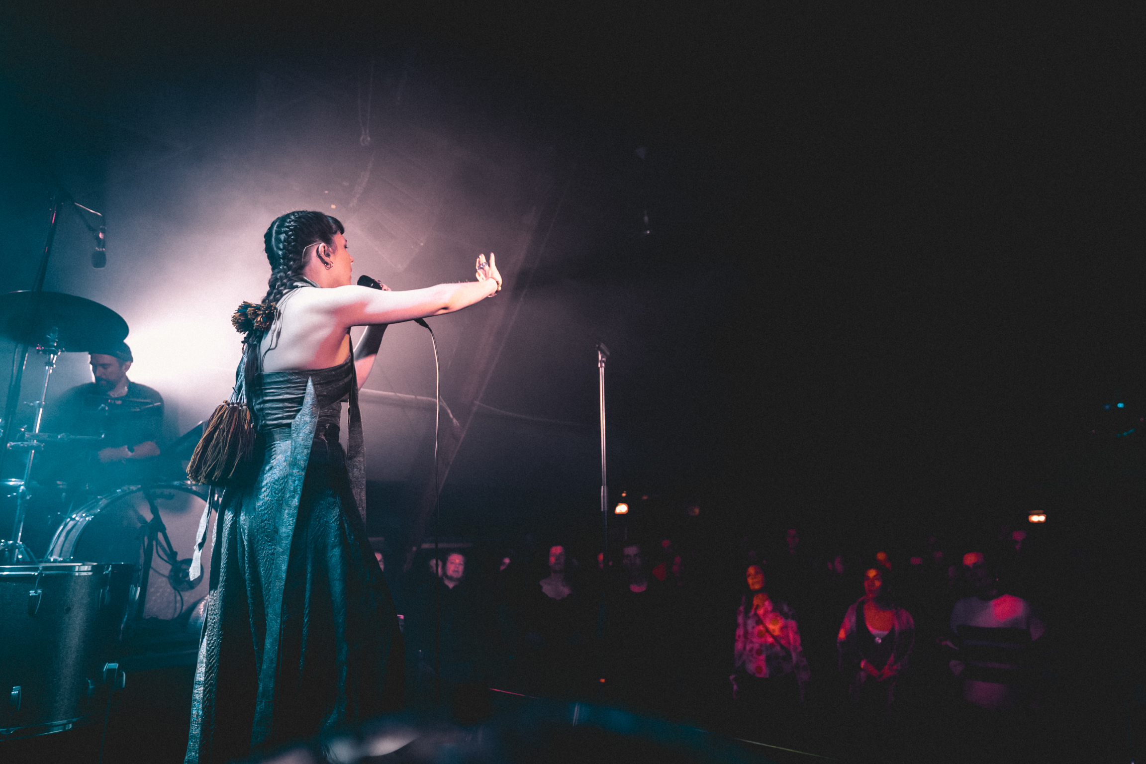 Female performer on stage with arm extended towards audience, backlit by bright white stage light against dark venue background.