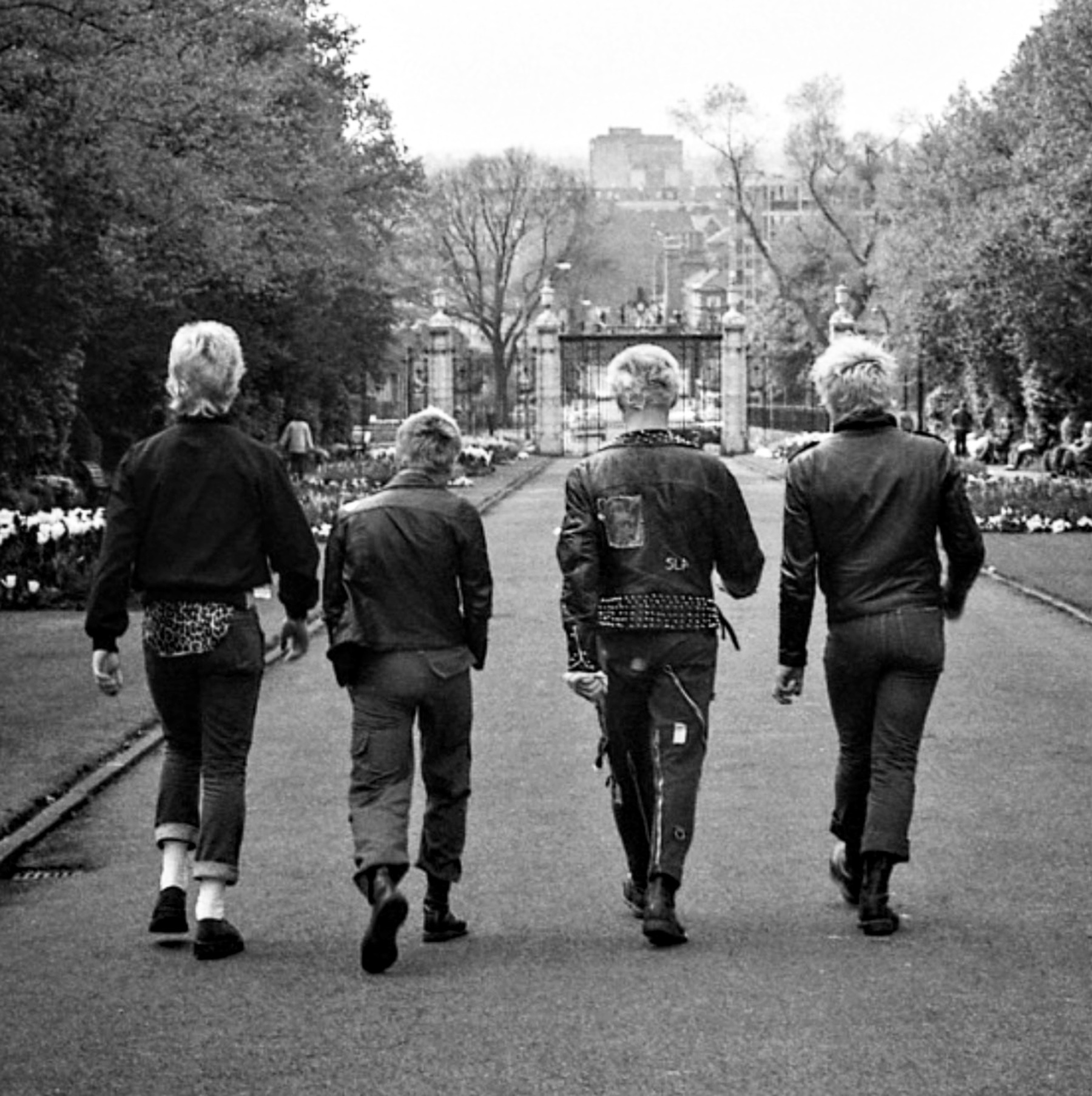 Four elderly men walking away from camera down tree-lined street towards ornate gates, black and white photograph.