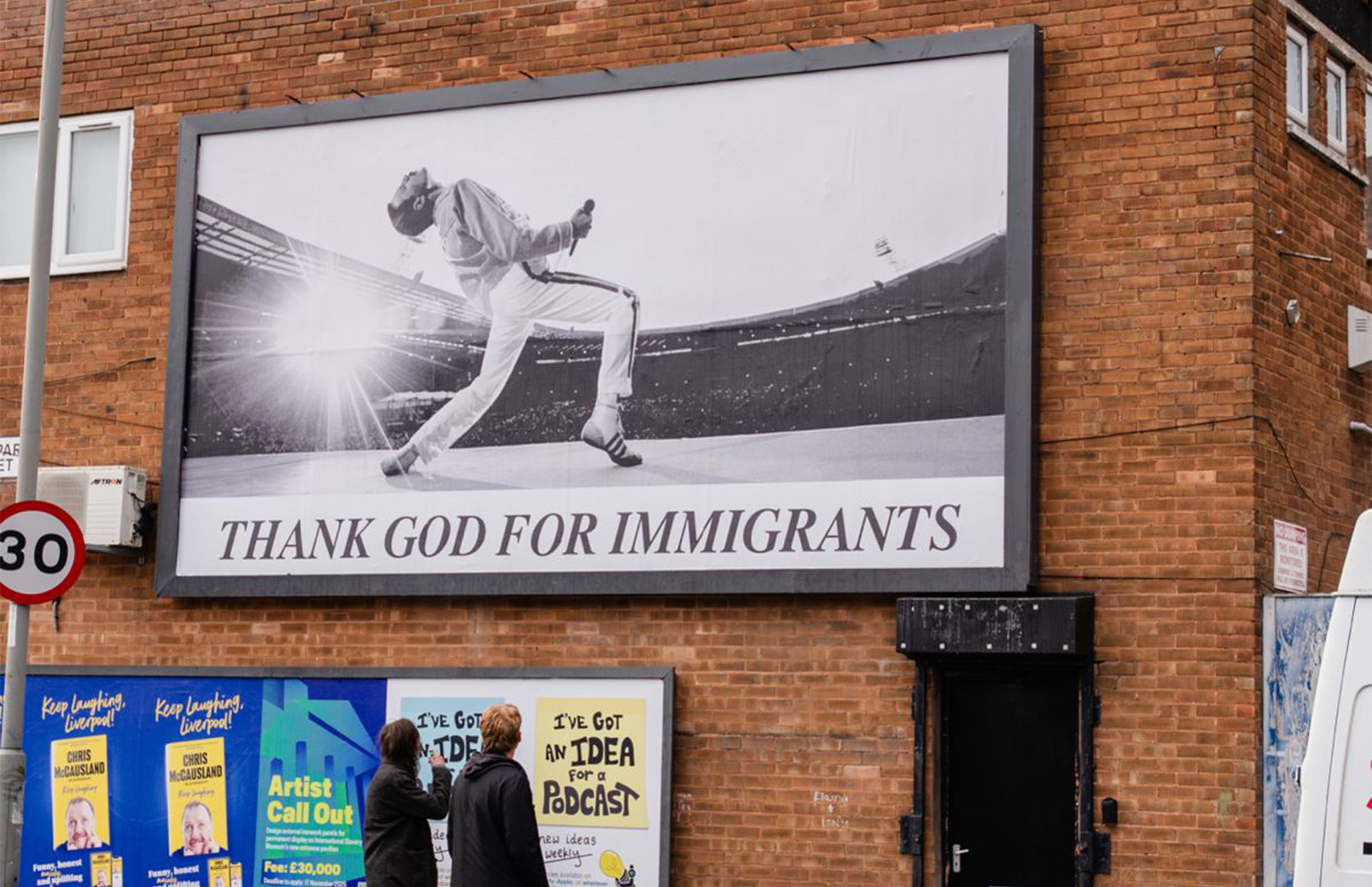 Large billboard on brick wall showing Freddie Mercury performing with text "THANK GOD FOR IMMIGRANTS" below smaller posters.