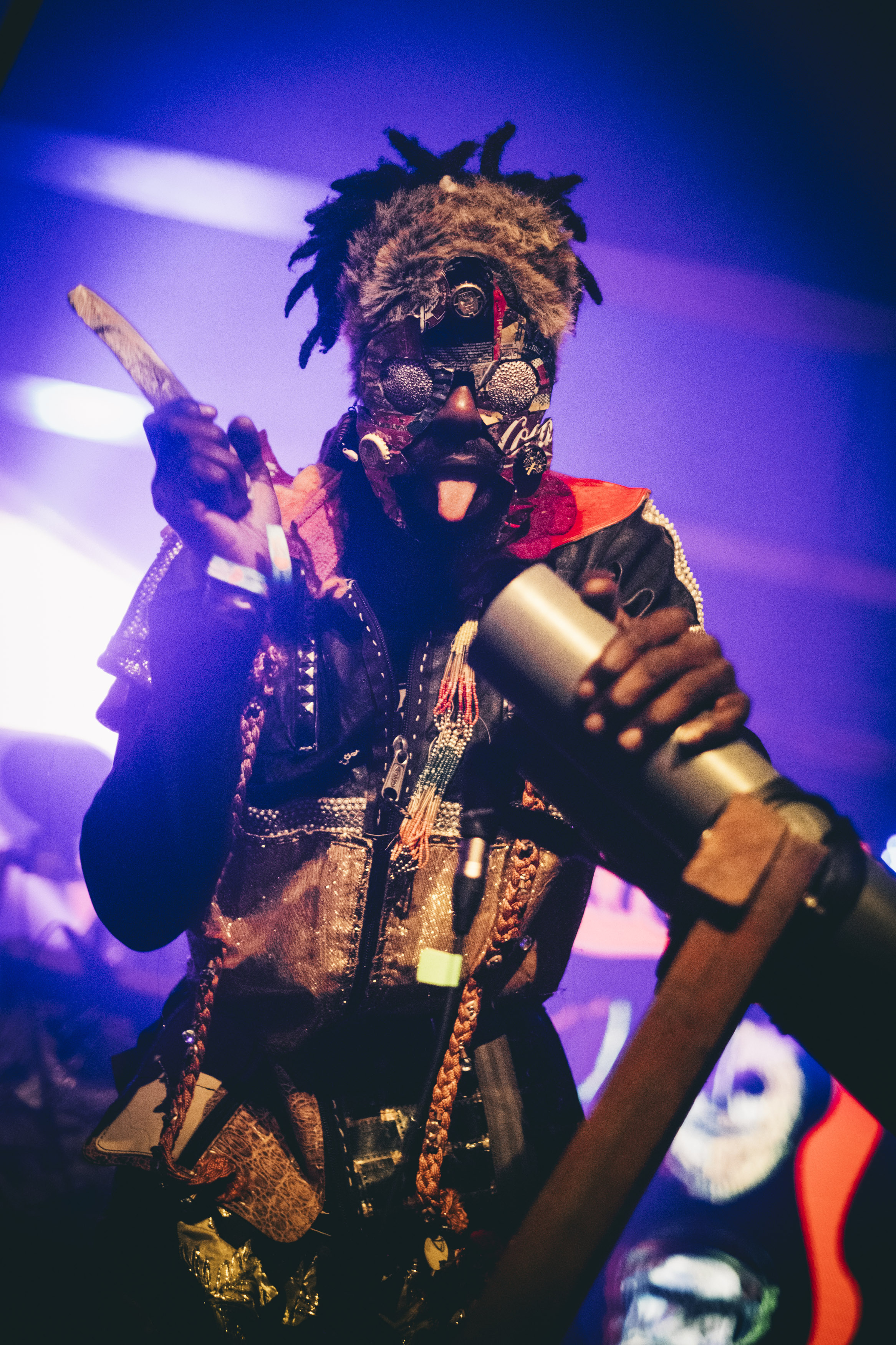 A figure wearing an elaborate, colourful costume with feathers and beaded accessories, holding a microphone while performing on stage under vibrant blue and purple lighting.