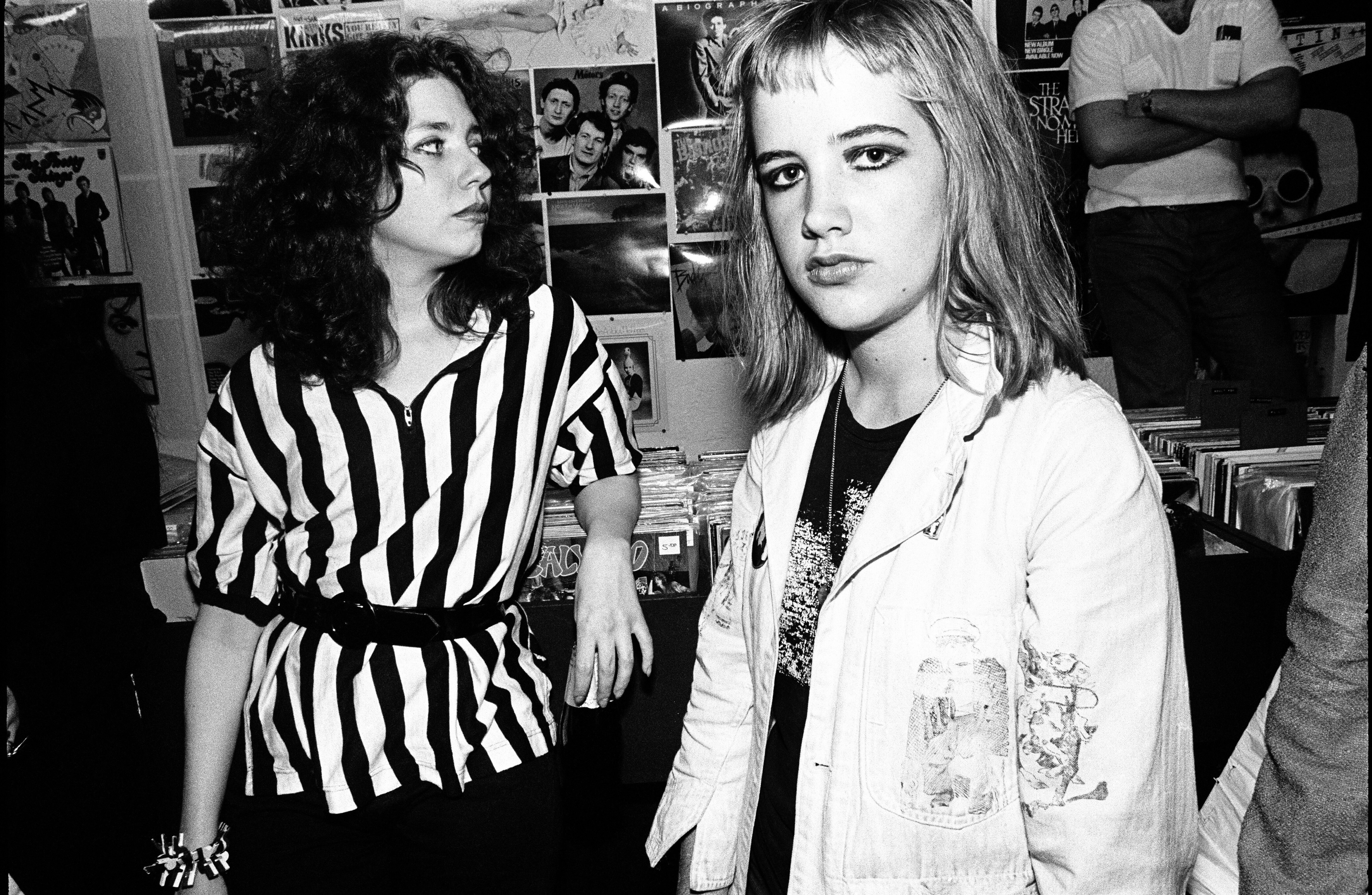 Two women, one wearing a striped dress, the other a white jacket, pose against a backdrop of posters.