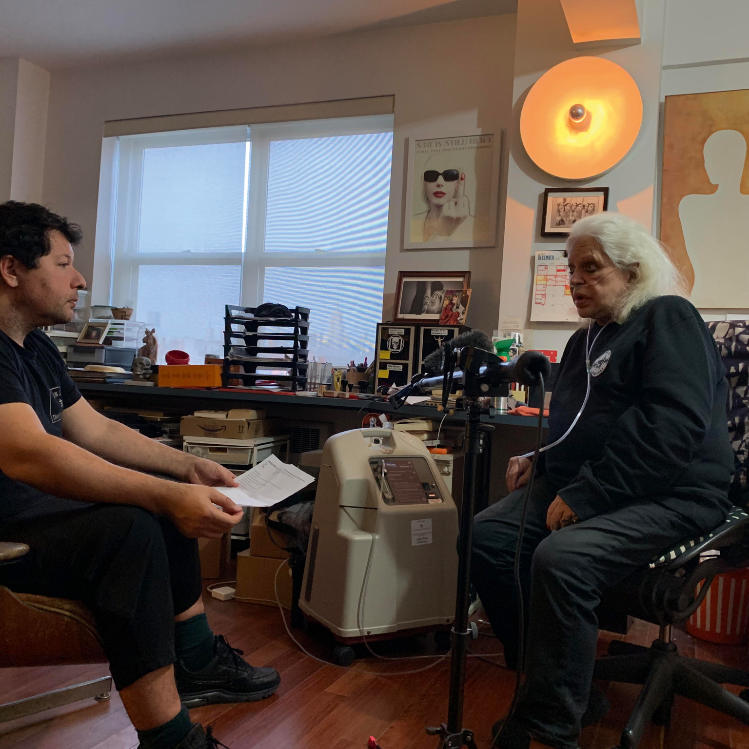 Two men in cluttered office space with large window, orange lamp overhead, one seated at desk with papers, other in chair nearby.