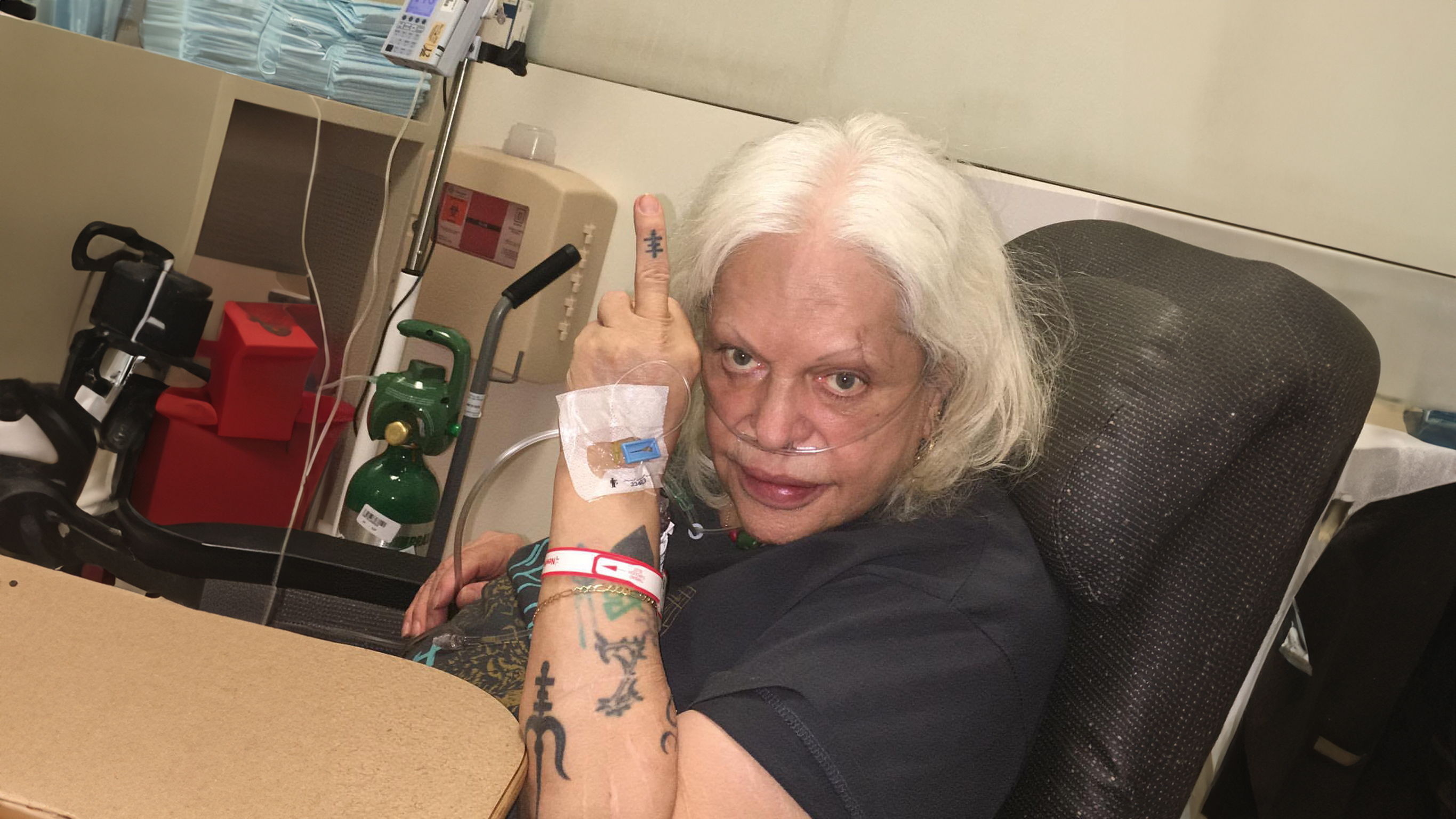 Elderly woman with white hair giving middle finger gesture whilst sitting in chair, wearing hospital bracelet in clinical setting.
