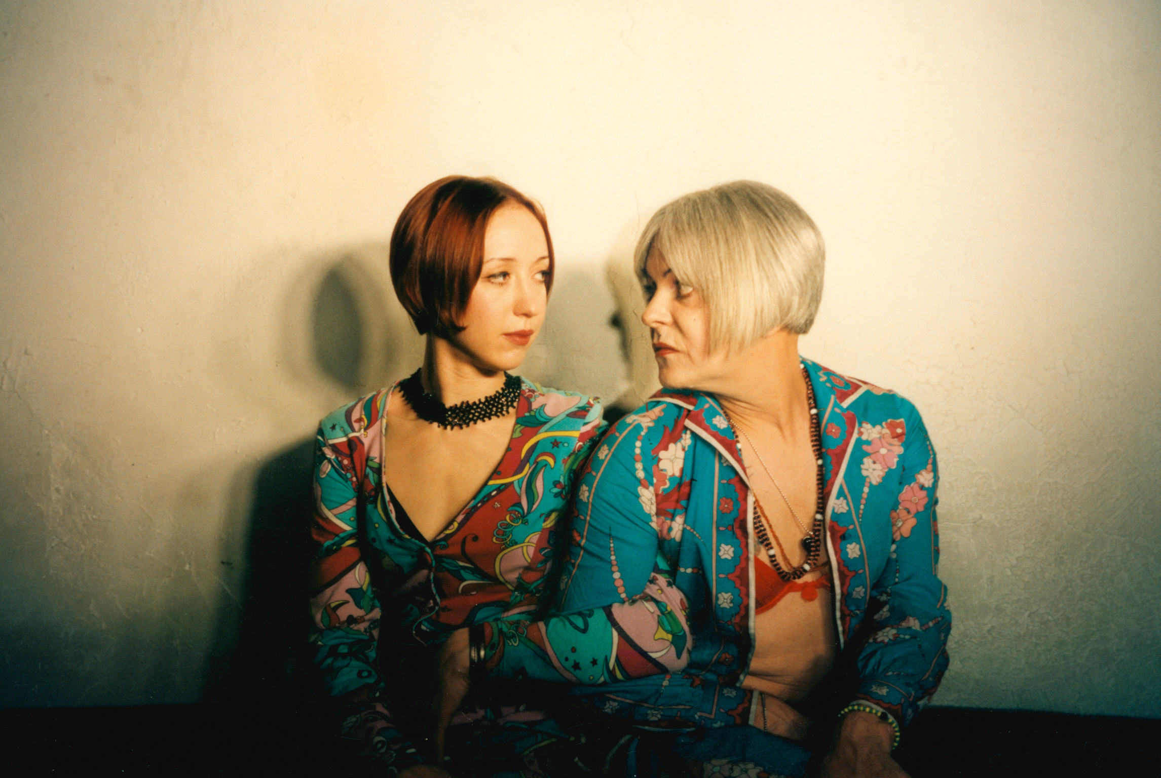 Two people in matching turquoise floral shirts facing each other against cream wall, one with dark bob haircut, other with platinum blonde hair.