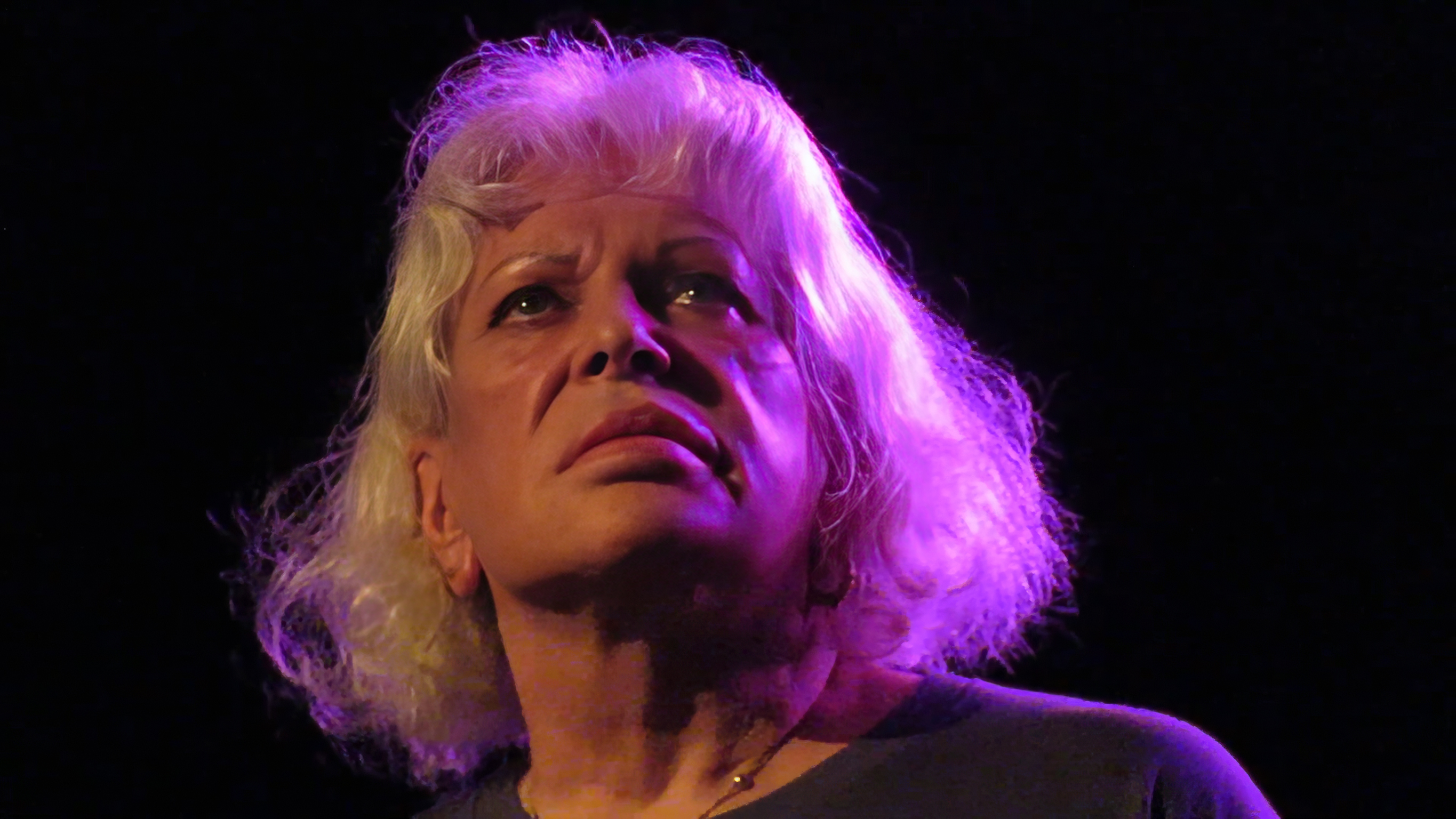 Elderly woman with blonde hair under purple stage lighting against black background, looking upward.