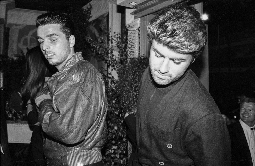Two young men in 1980s clothing walking together, one in patterned jacket, one in dark top, black and white photograph.