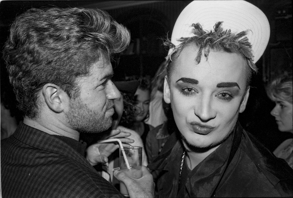 Black and white image of two people at a social gathering. Man with curly hair on left, person with short spiky hair and white beret on right.