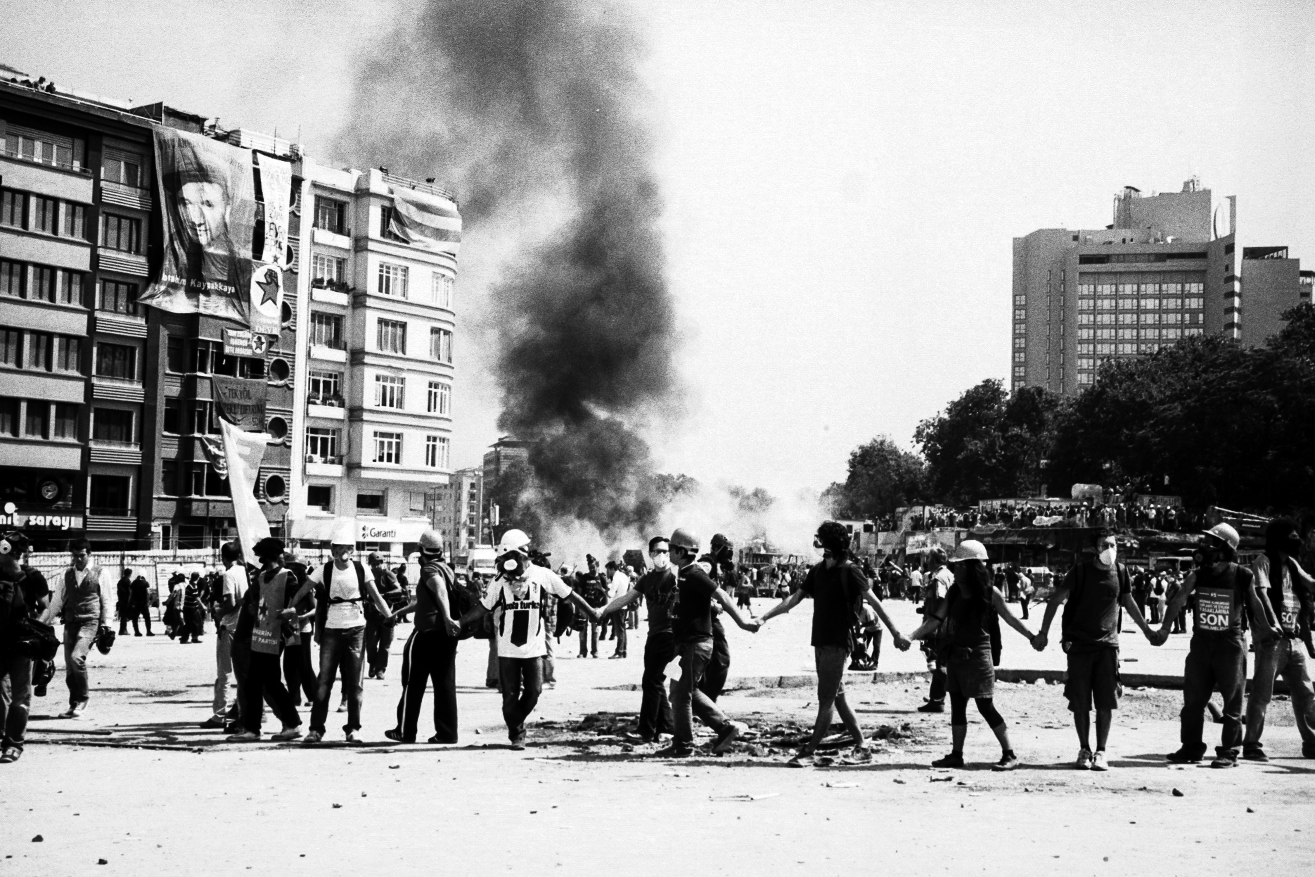 Outdoor scene of a large crowd gathered in a city street, with smoke and flames visible in the background.