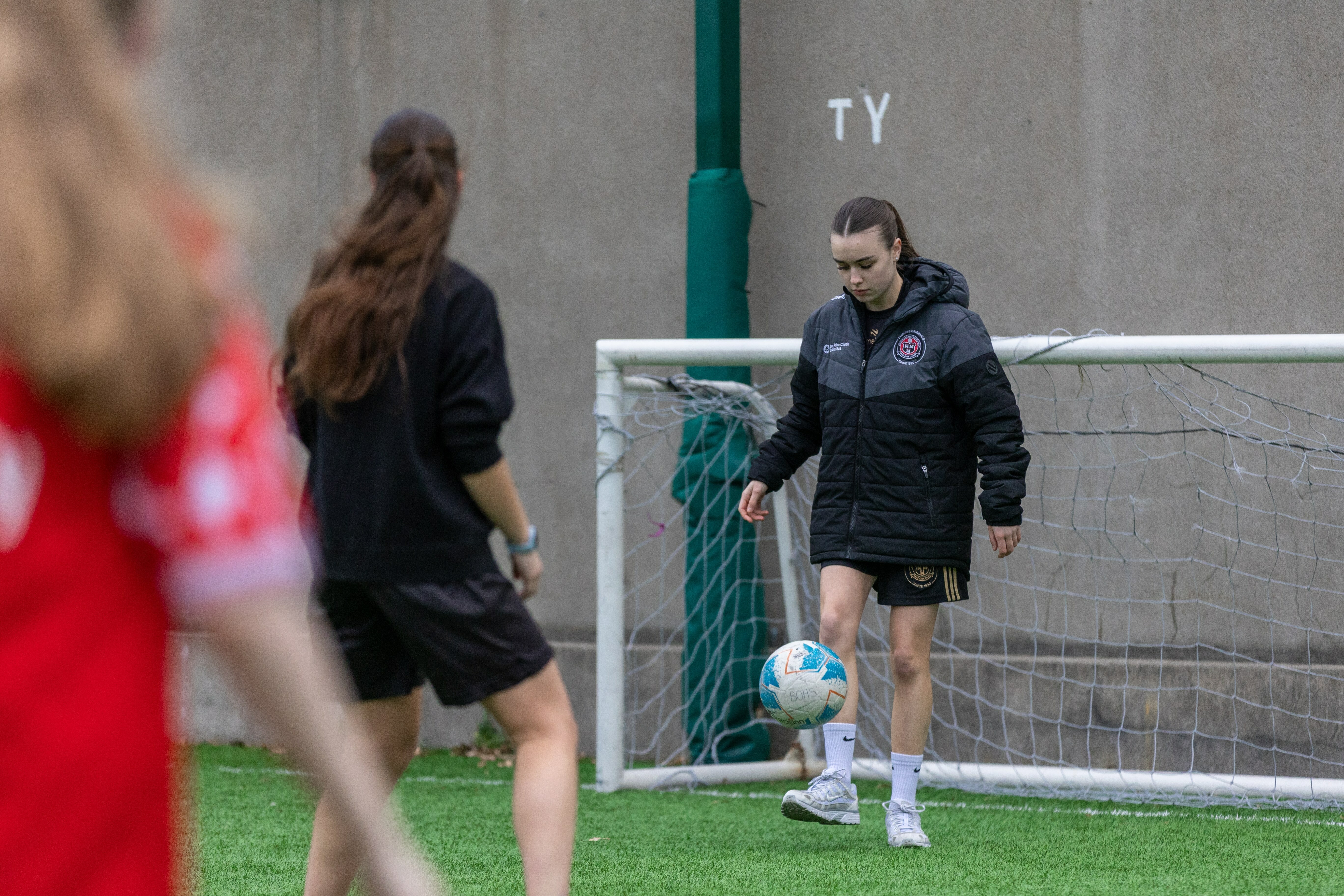 Two people playing football on a pitch with a goal post and 'Ty' signage visible in the background.