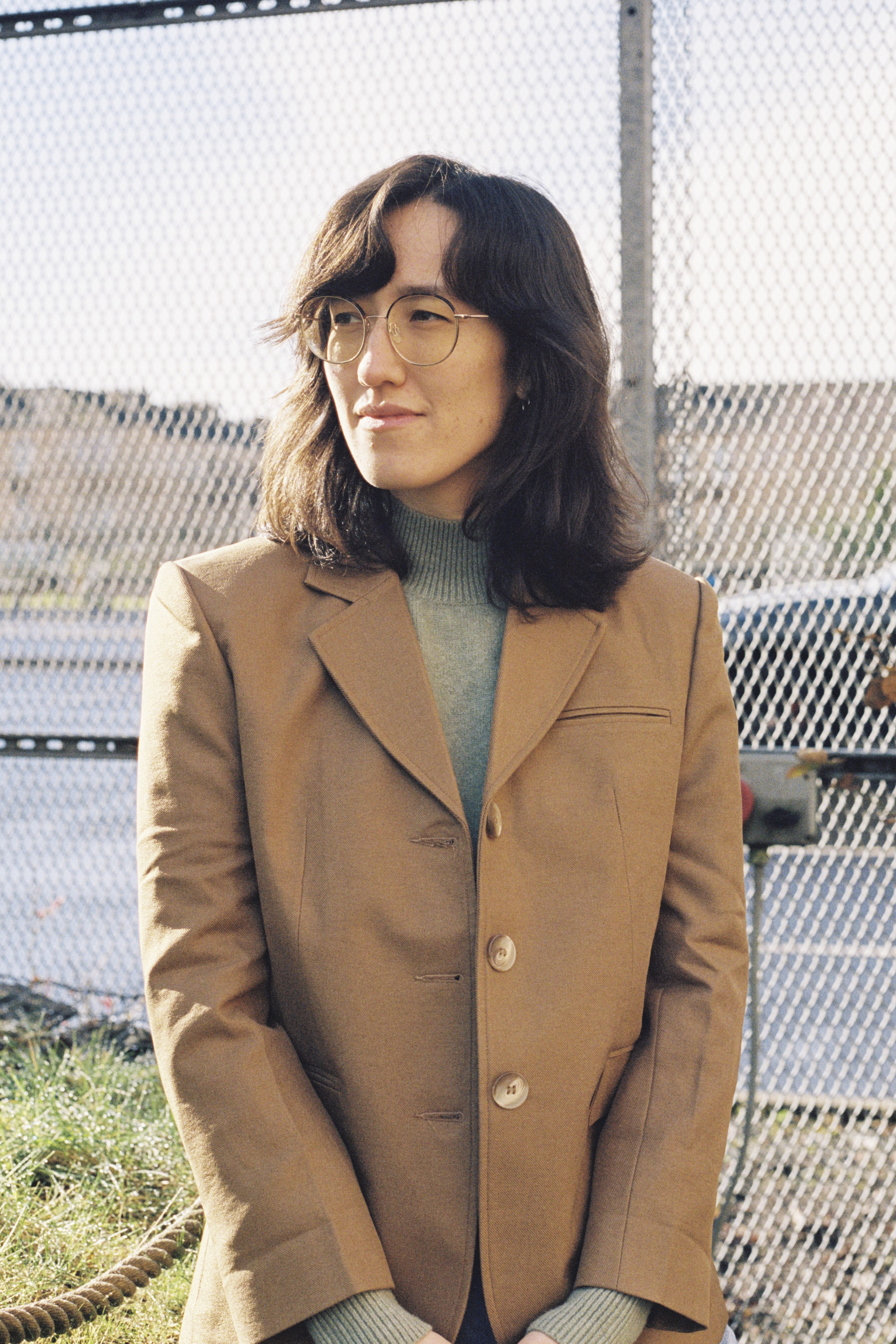 A woman with dark hair and glasses wearing a beige coat and standing in front of a wire mesh fence.